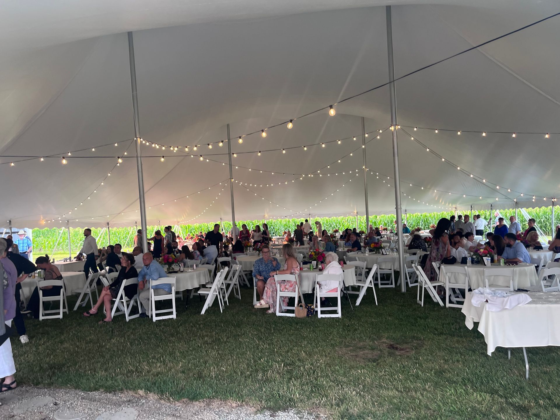 Guests seated at tables under a white tent, string lights overhead, outdoor gathering.