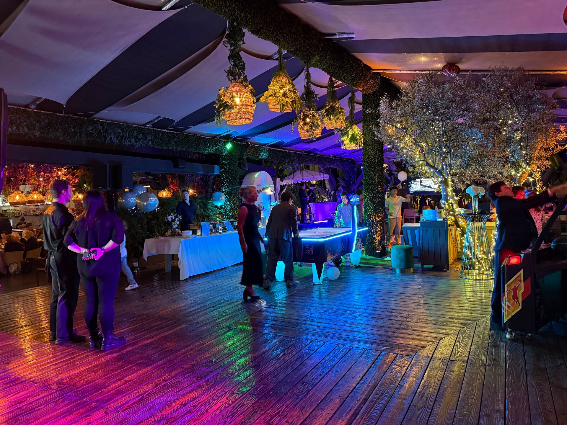 People in a brightly lit outdoor space with a bar, wooden floor, and colorful lights.