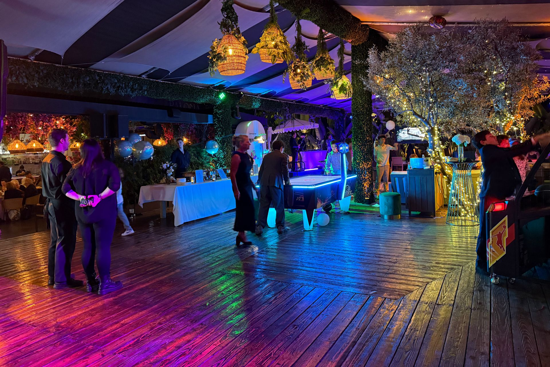 An outdoor party with colorful lights, people gathered around a pool table and tables.