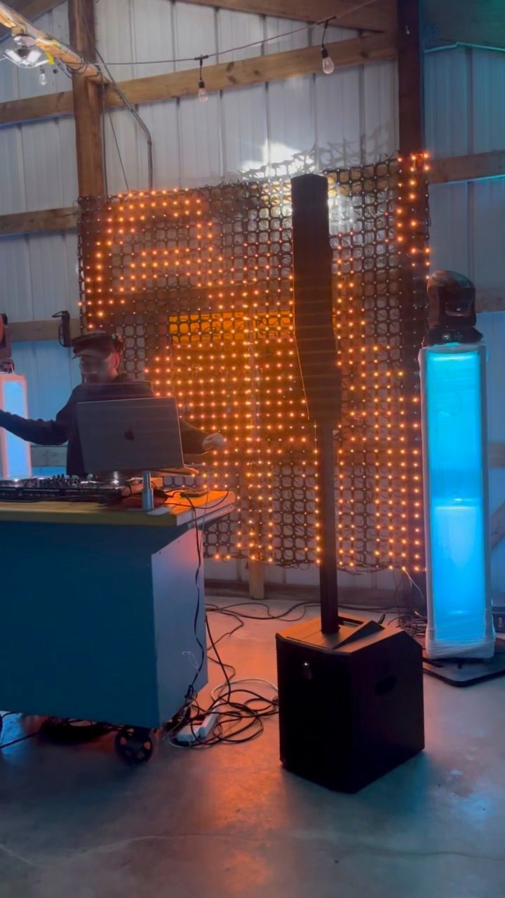 DJ at a party. Lights illuminate a backdrop. DJ console and speakers are present.
