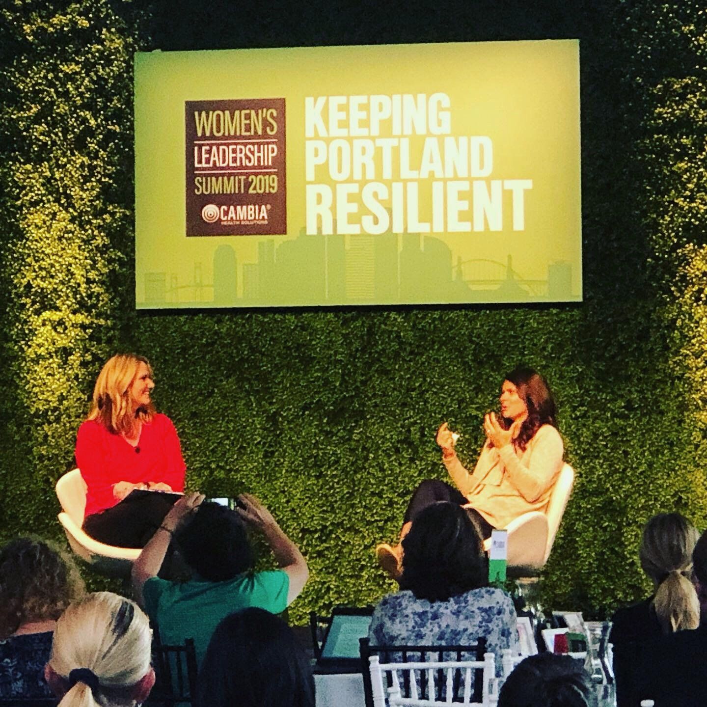 Two women speaking on stage at the Women's Leadership Summit 2019 in Portland. Audience watching.