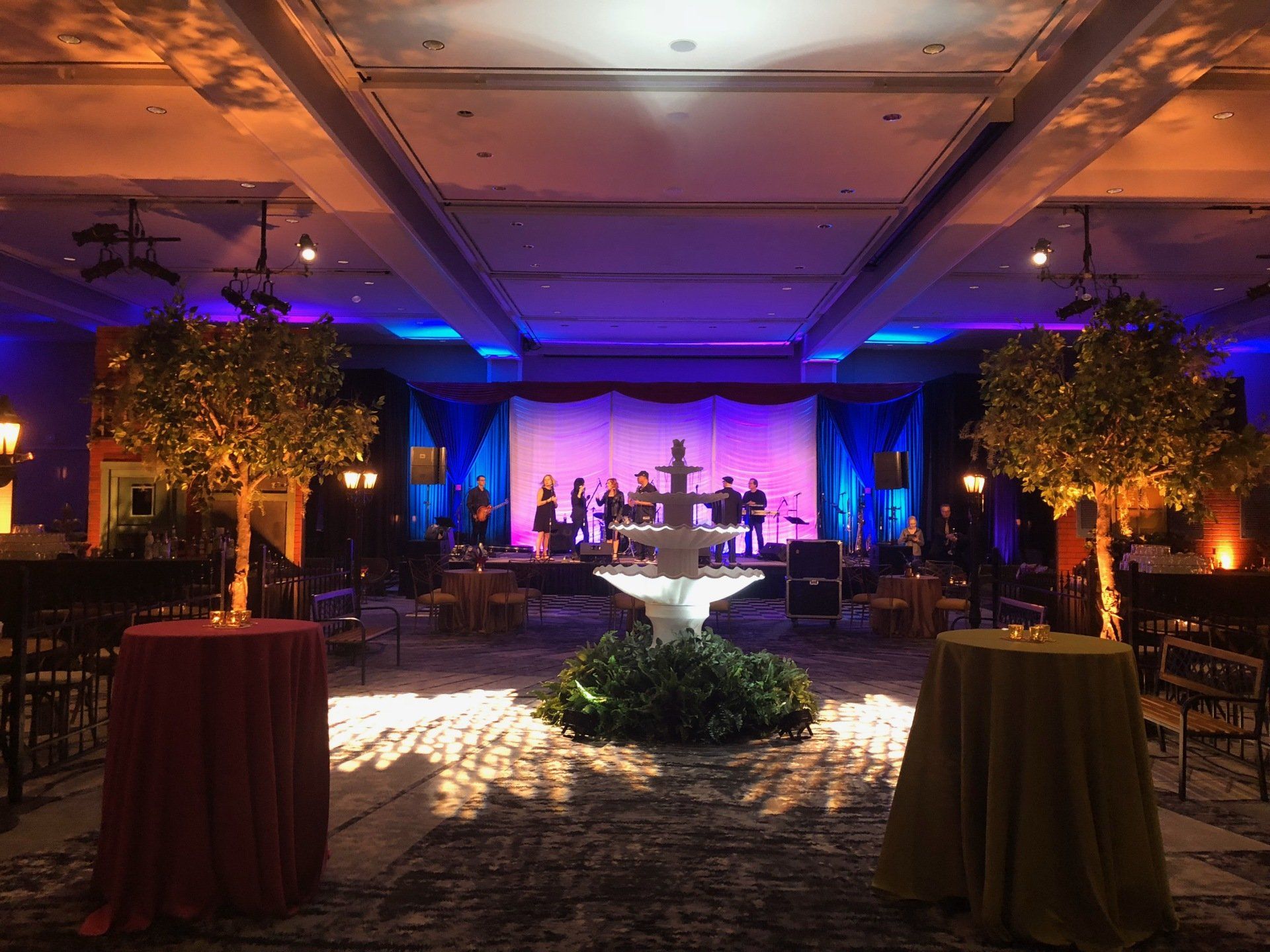 Event space with band on stage, fountain centerpiece, decorated trees, tables with colorful linens, and blue and purple lighting.