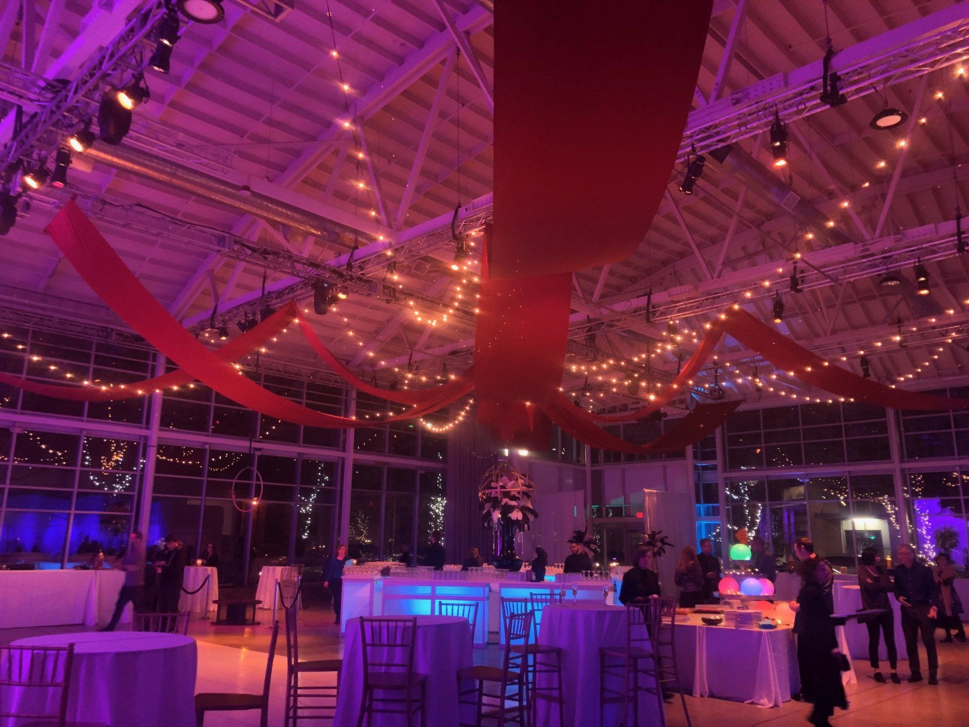 Event hall with white tables, chairs, and red draping; strings of lights and purple lighting.