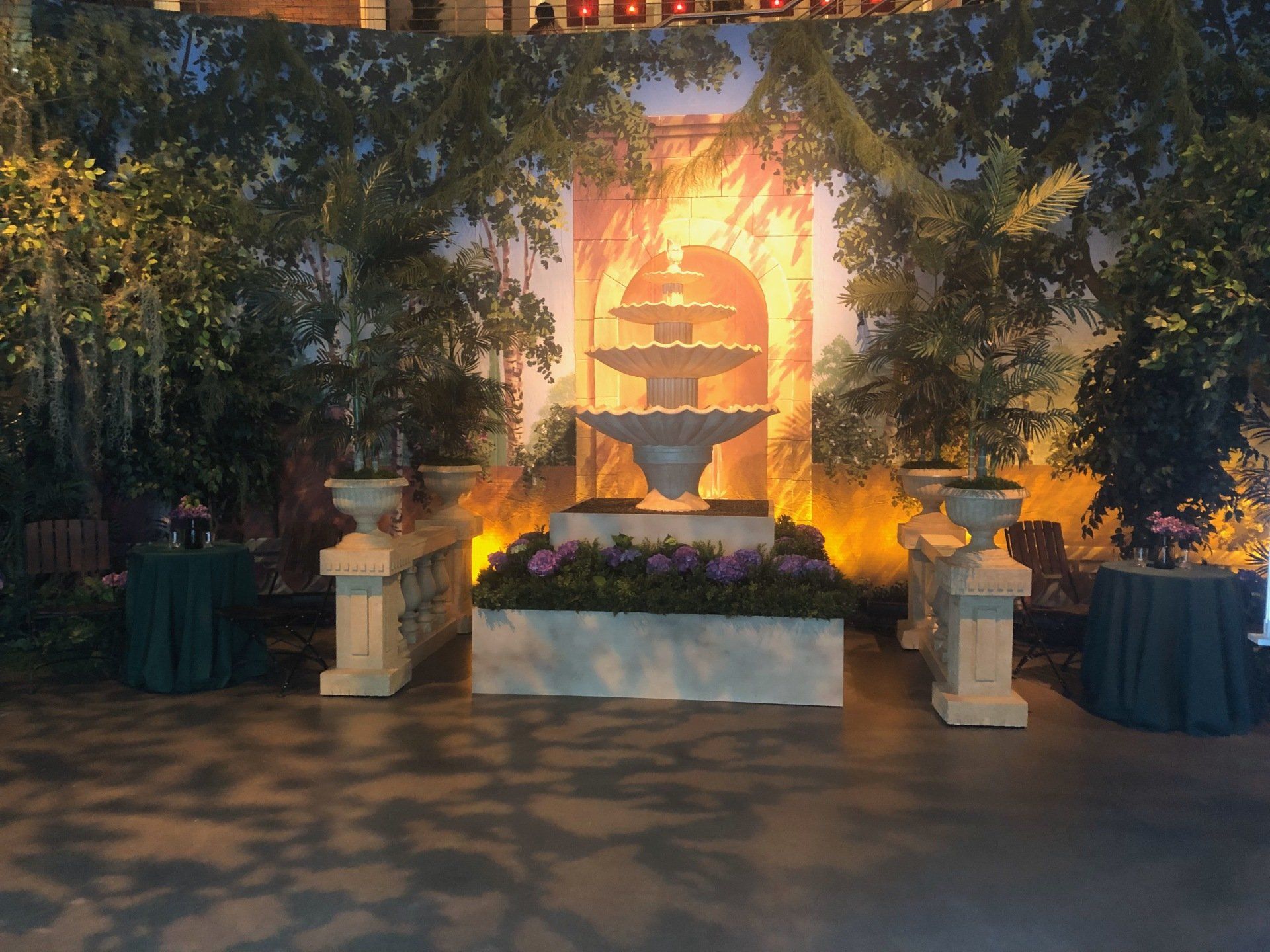 Fountain scene with a tiered fountain, flanked by urns and greenery, against a painted backdrop of a building.