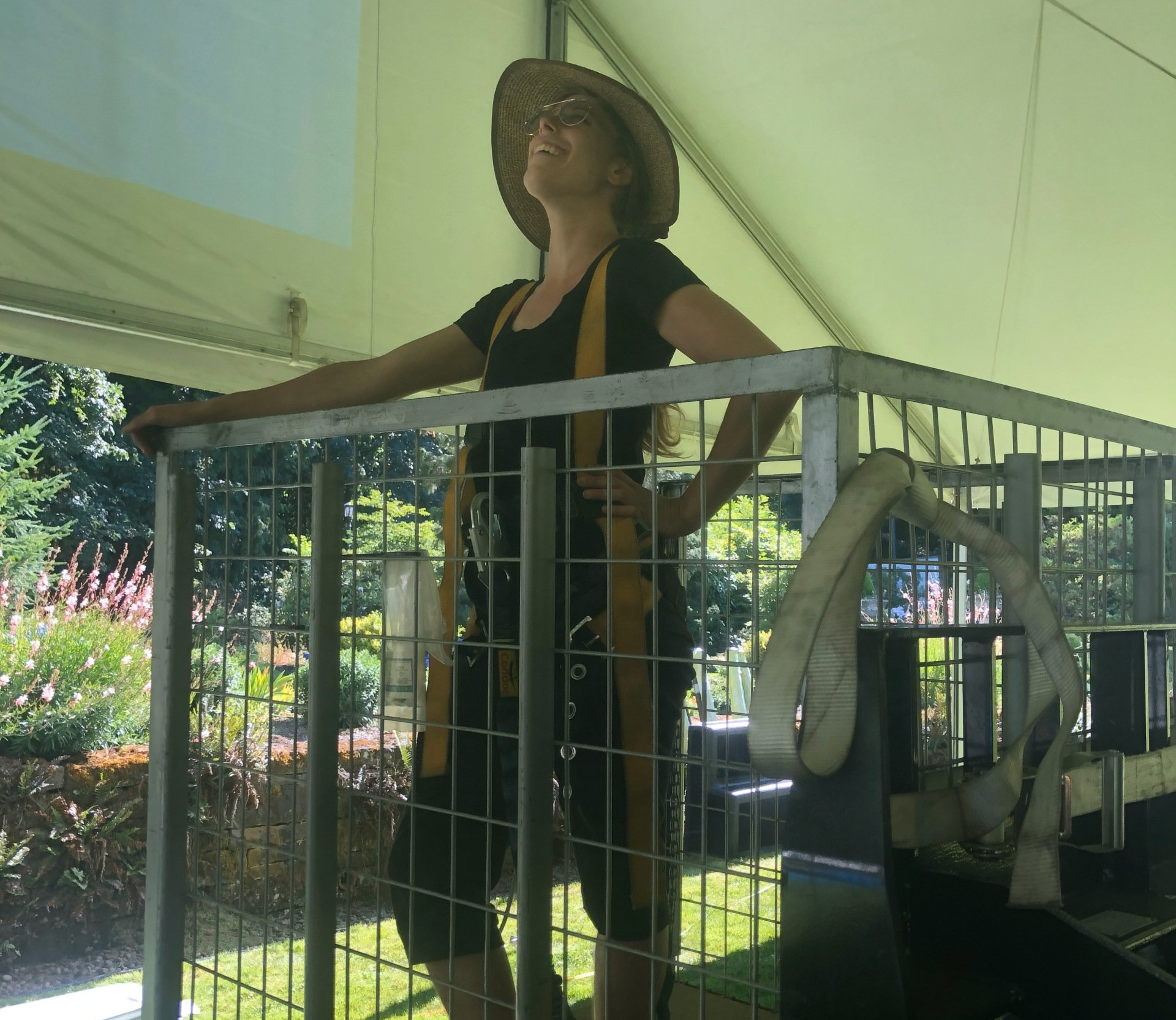 Woman in straw hat smiles, leaning on a metal cage structure. Outdoors, sunny, trees in background.