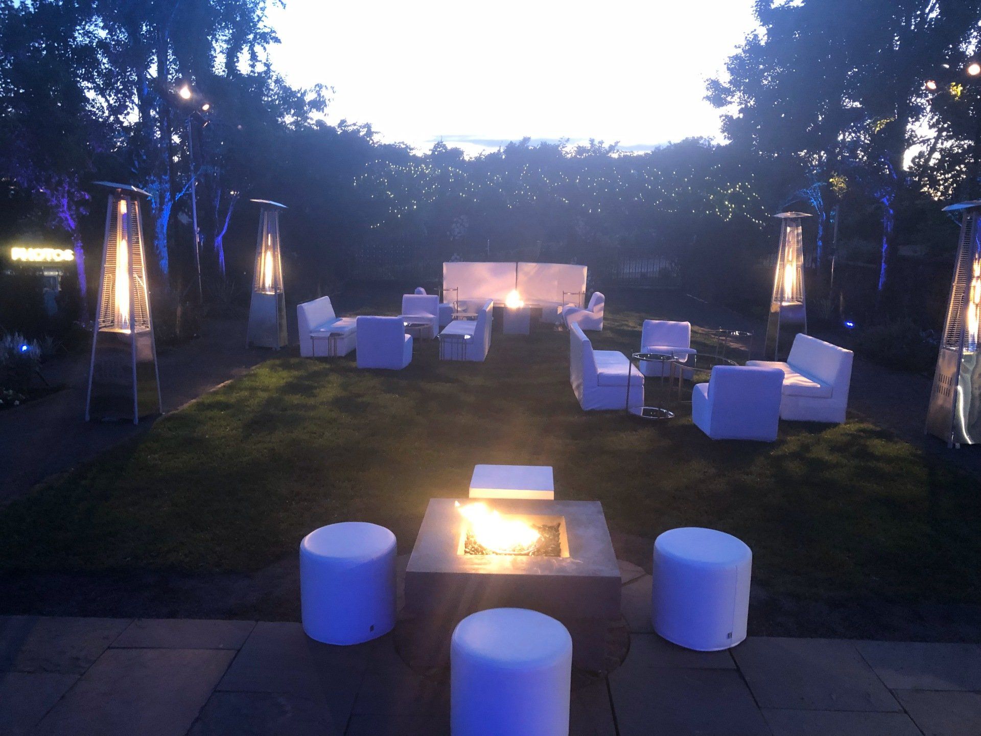 Outdoor lounge with white furniture, fire pits, and lit trees at dusk.