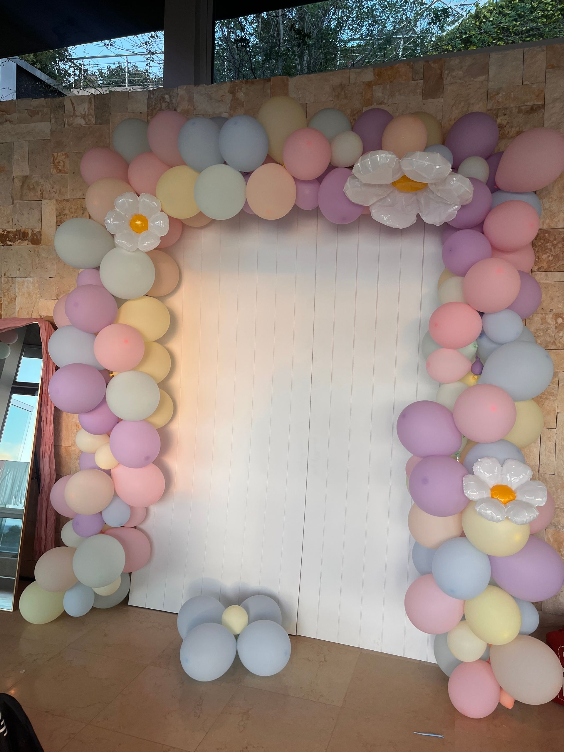 Archway of pastel balloons, with daisy accents, framing a blank white backdrop, set indoors.