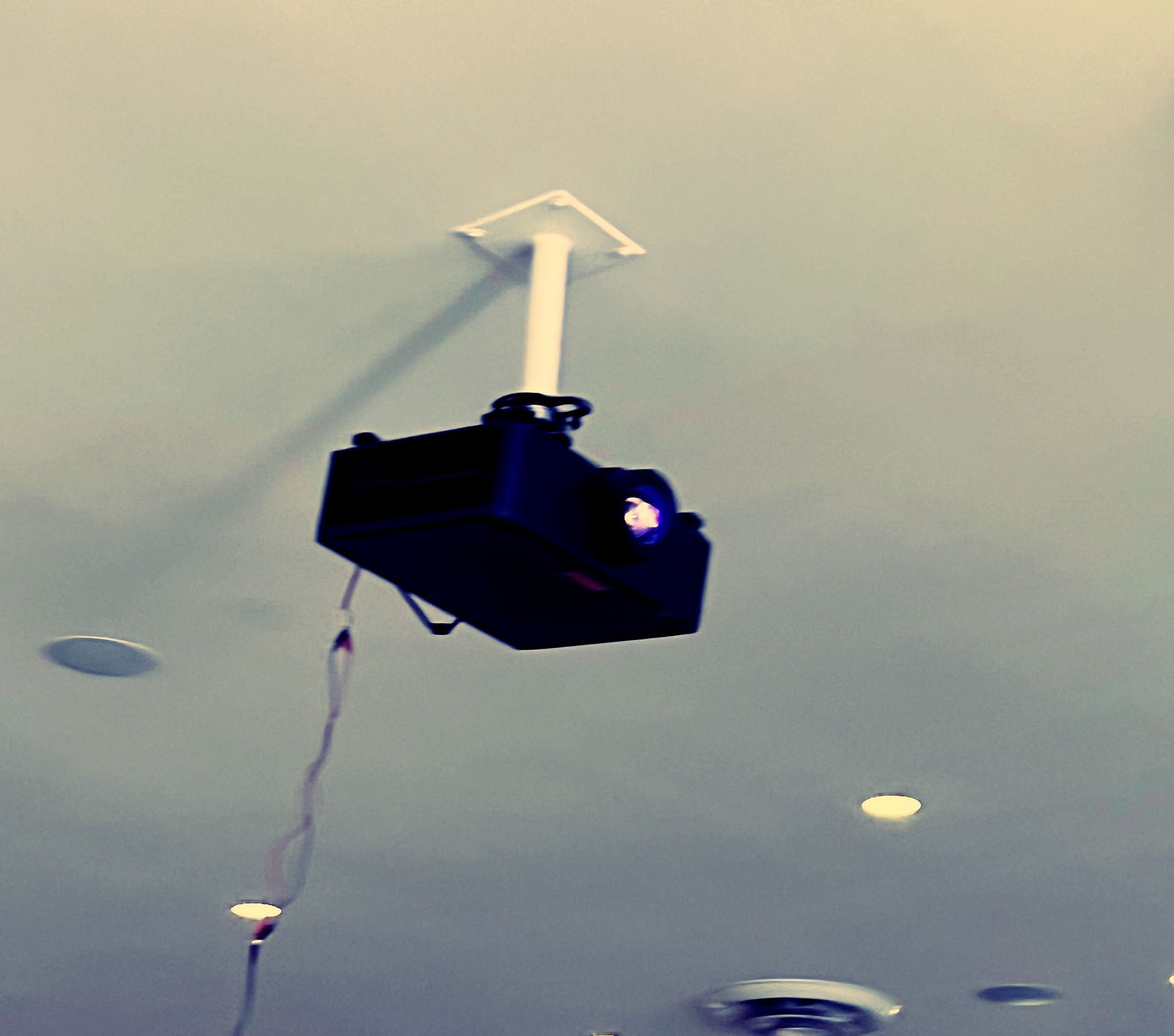 Projector suspended from a white ceiling, with visible cords and lighting fixtures below.