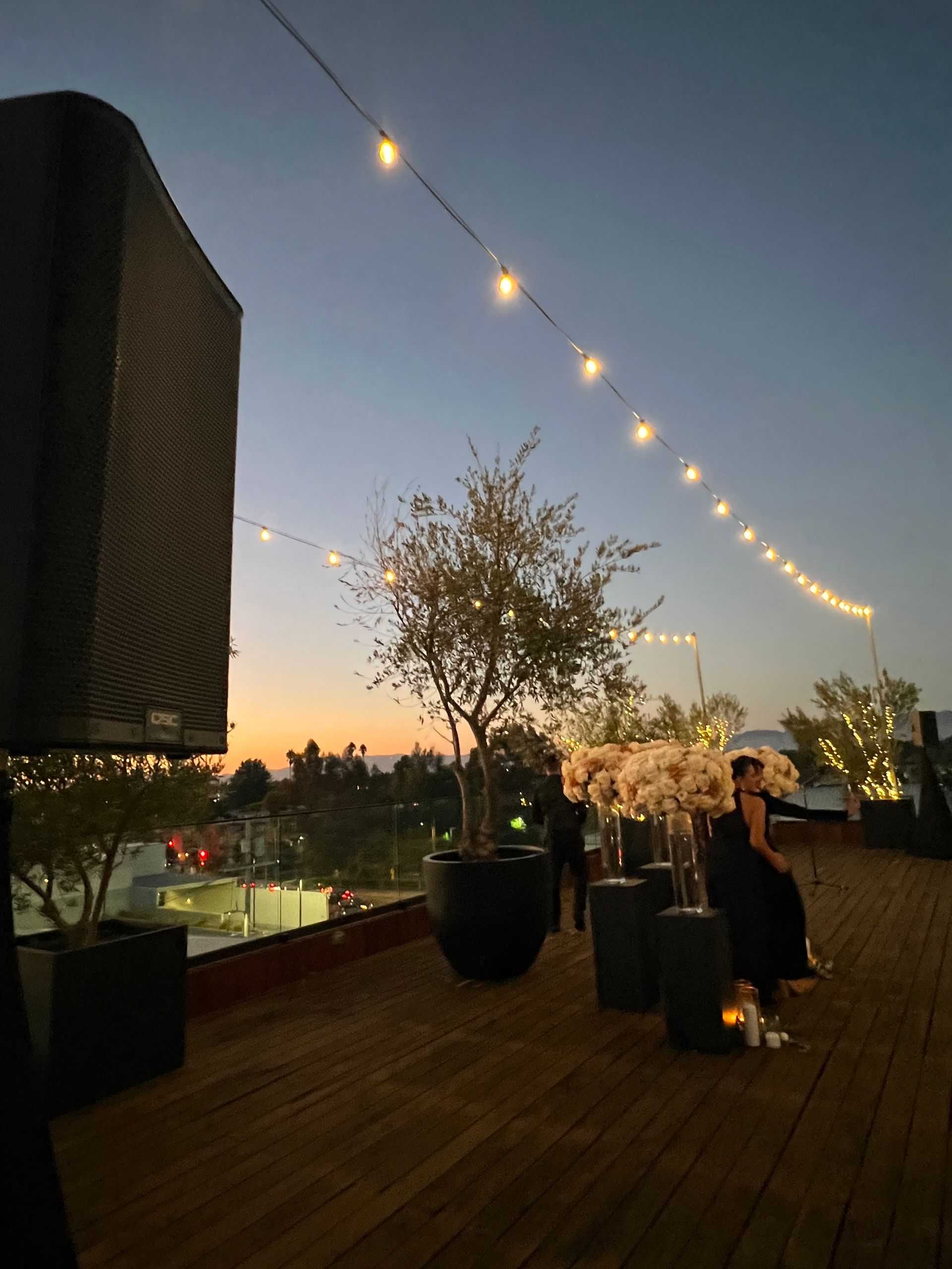 Rooftop event at dusk with string lights, trees, and people. Sunset colors in the sky and wooden deck.