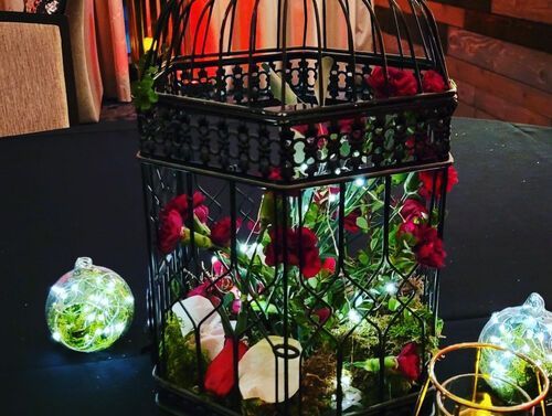 Black birdcage centerpiece filled with red roses, greenery, and fairy lights on a table.