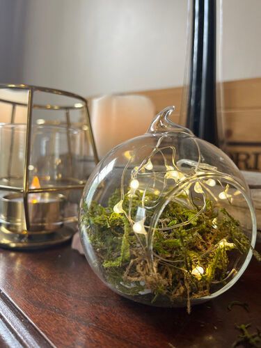 Glass orb terrarium with moss and fairy lights on a wooden surface next to a candle.