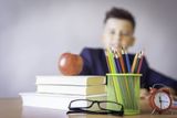 Books, pencils, apple, and clock on a desk with a blurred boy in the background.