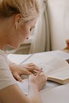 Blonde woman writing in notebook, studying a book, seated at a table, indoors.