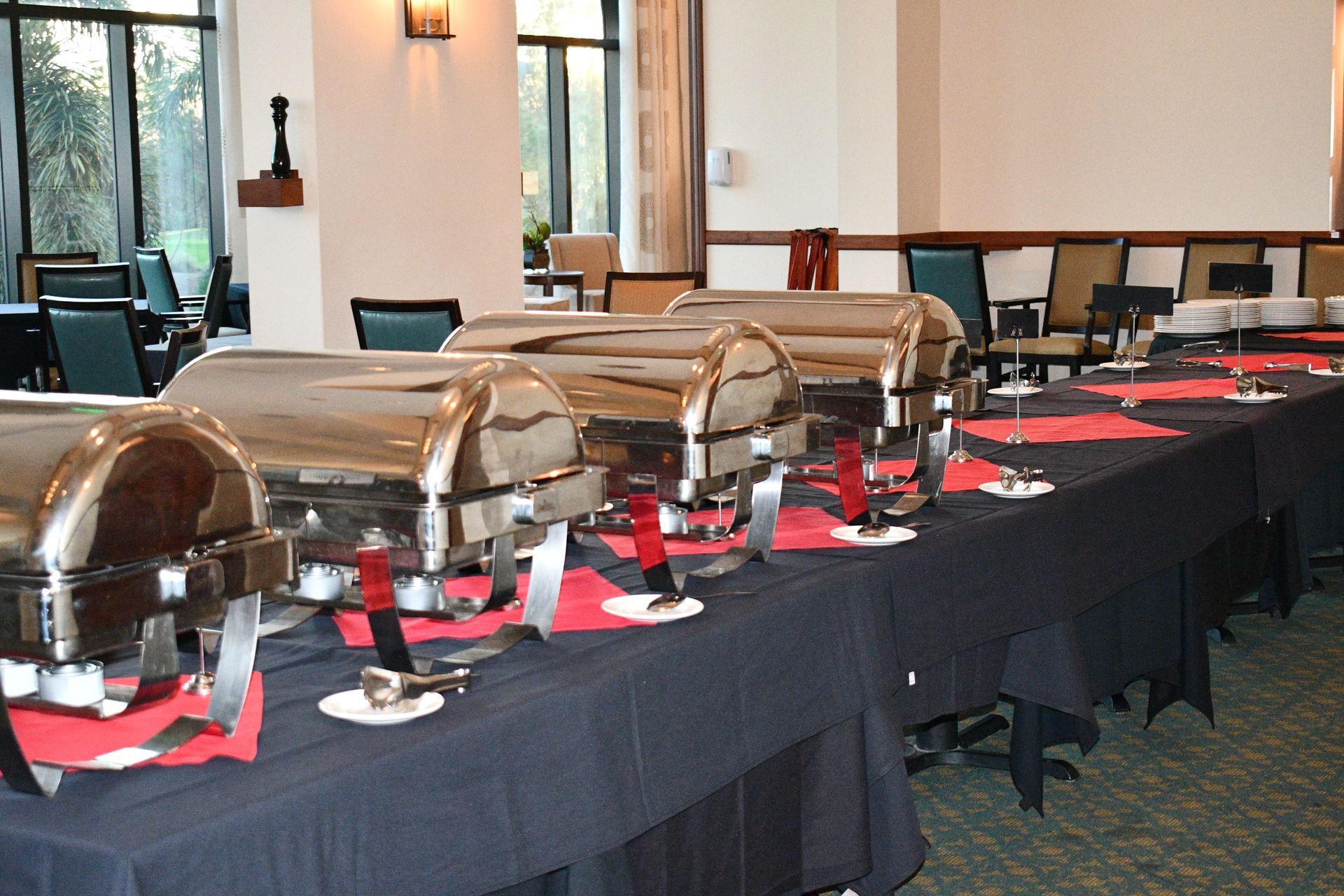 Buffet table set with silver chafing dishes and serving utensils in a room with a large window.