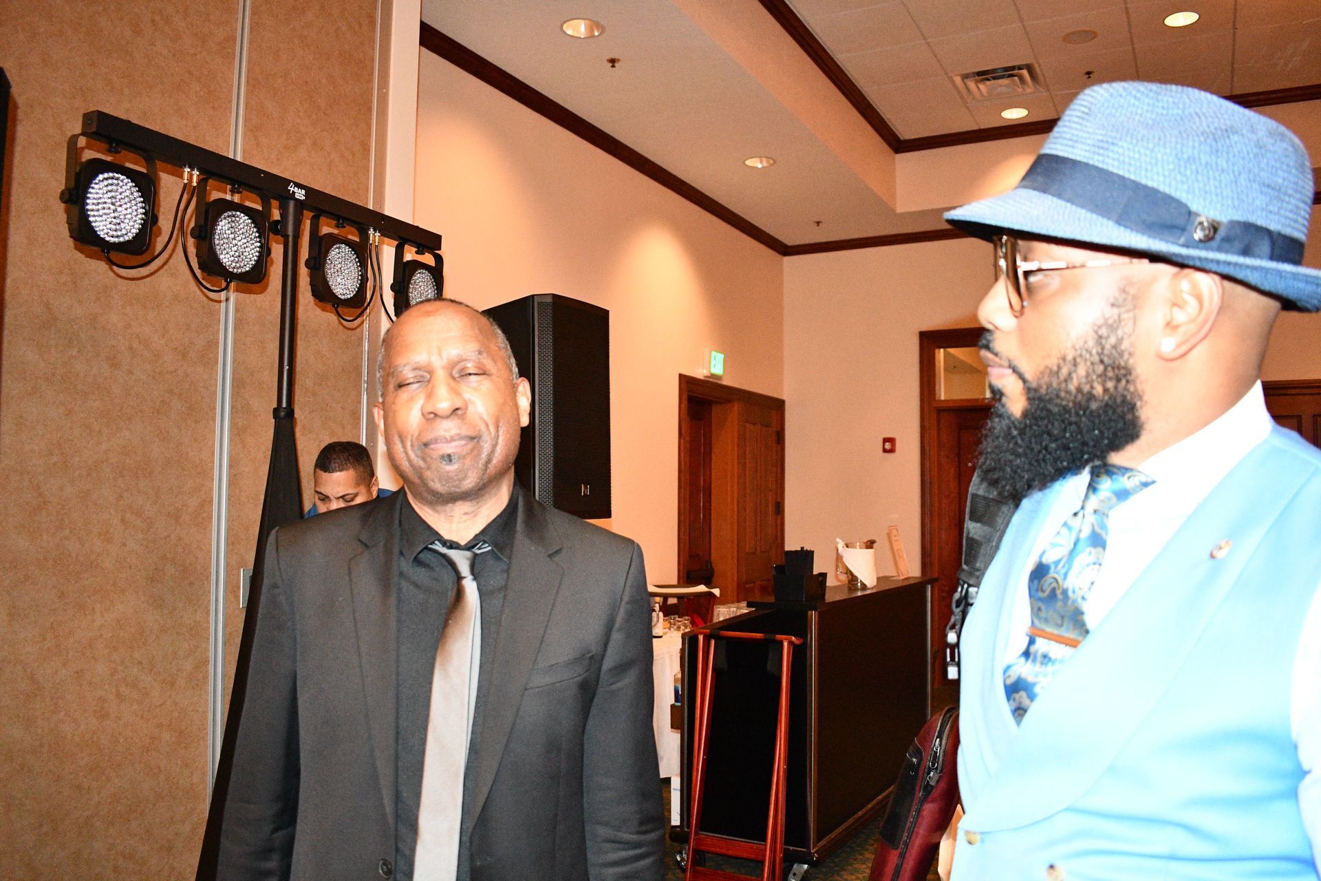 Two men in formal attire stand indoors, possibly at an event. Lighting rig on left.