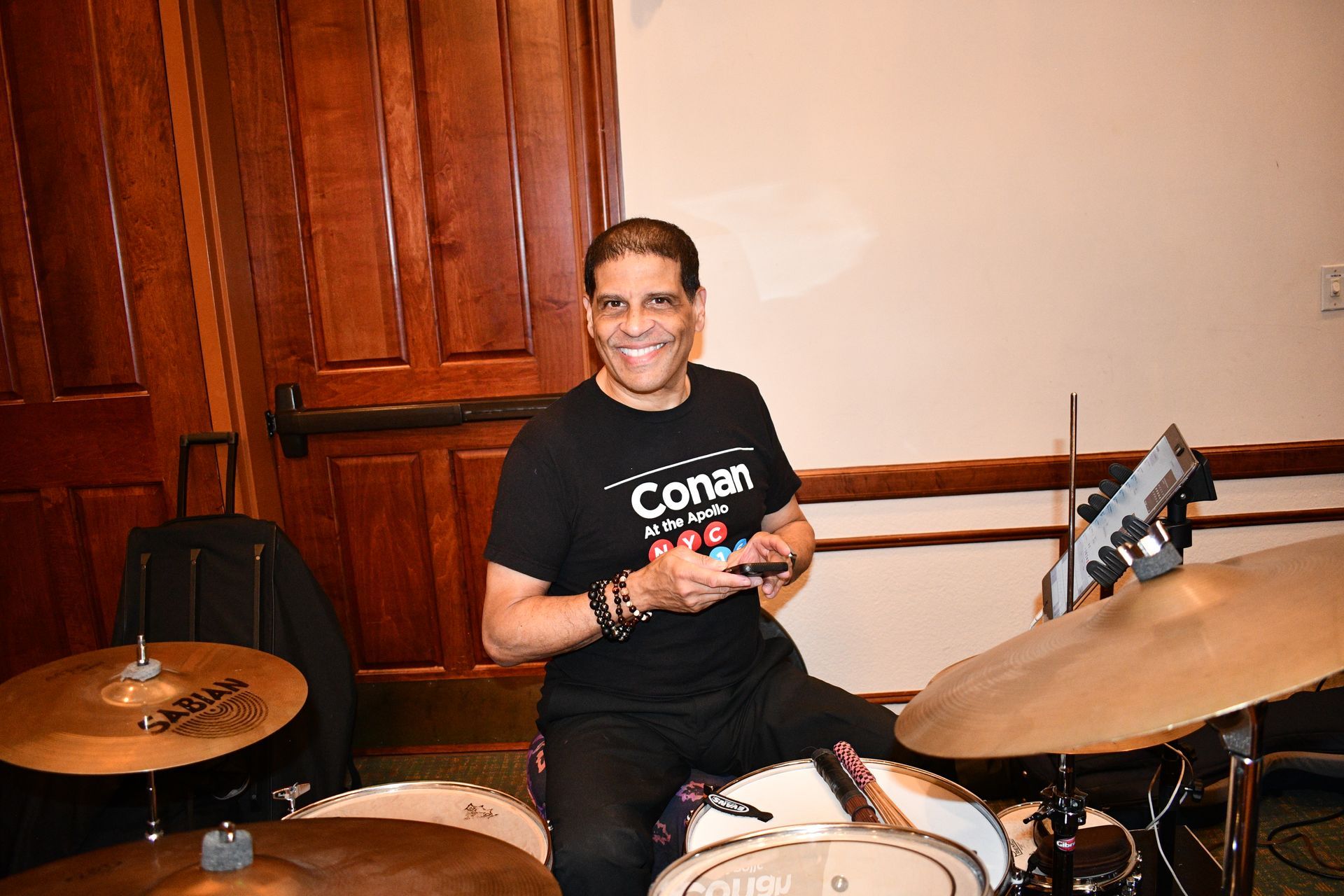 A smiling man in a Conan t-shirt sits at a drum set. Wooden background.