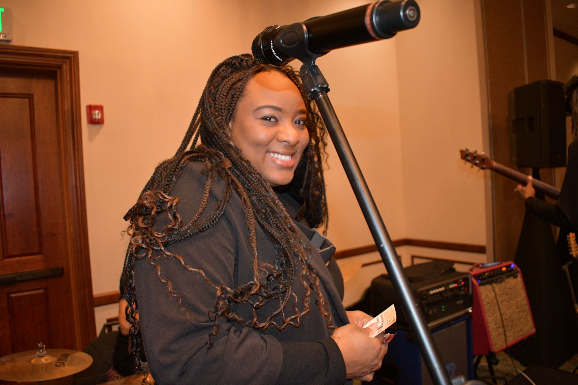 Woman smiling, holding cards, near microphone and band equipment. Indoors, warm lighting.