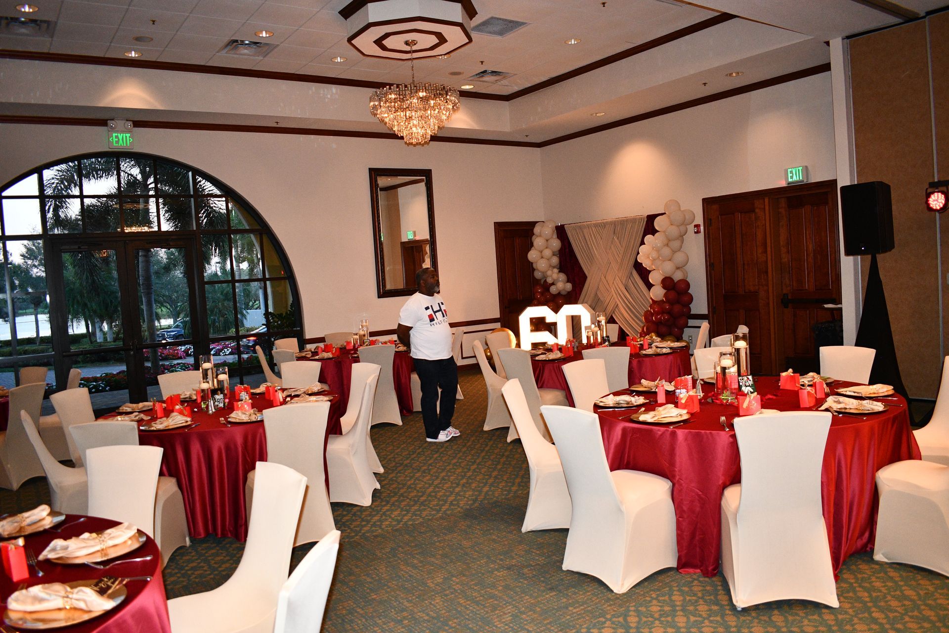 Banquet hall decorated for a celebration; round tables with red linens, white chairs, and floral centerpieces. A person stands near a table.
