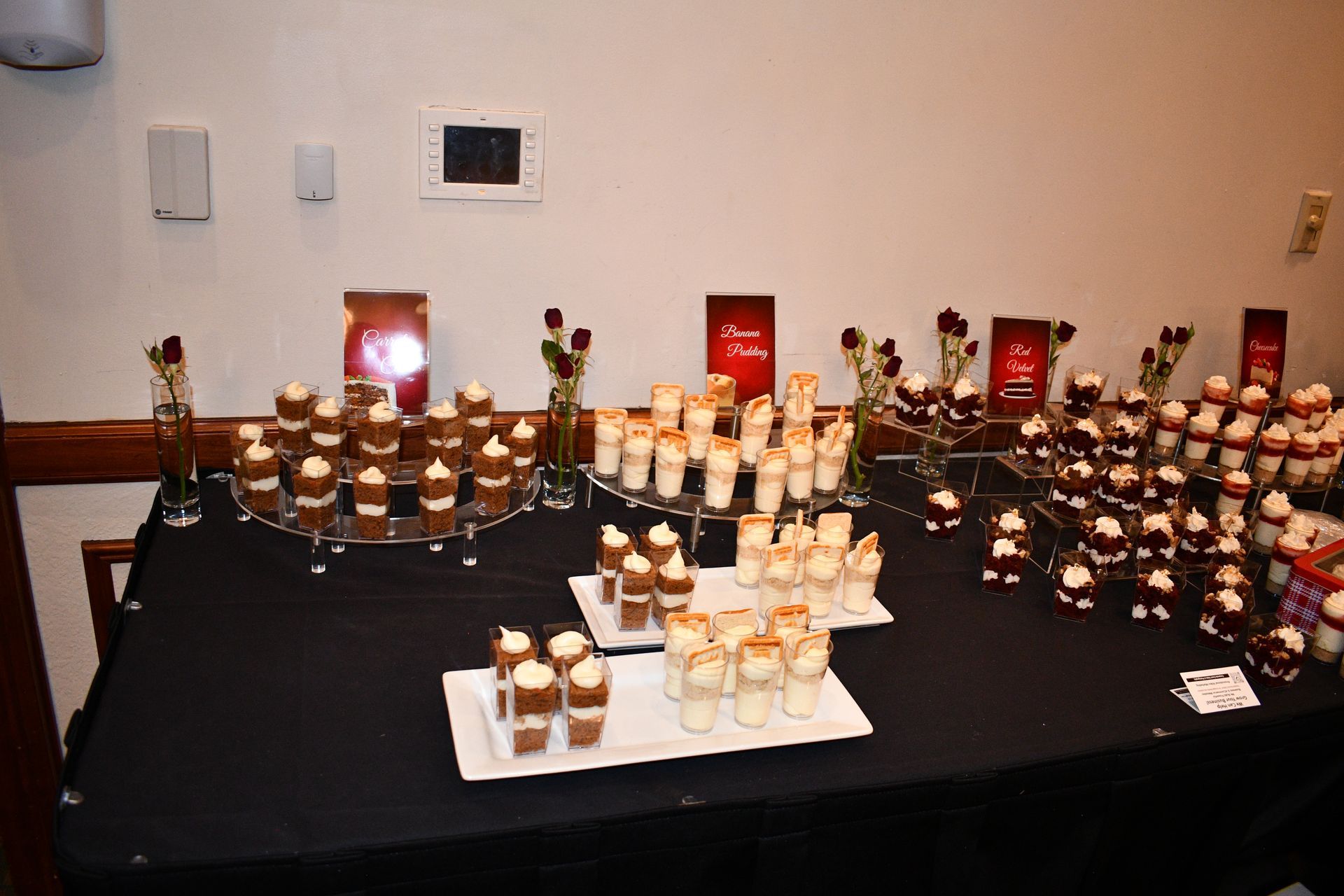 Dessert table with various layered treats on white trays and stands, black tablecloth, red roses, and two signs.