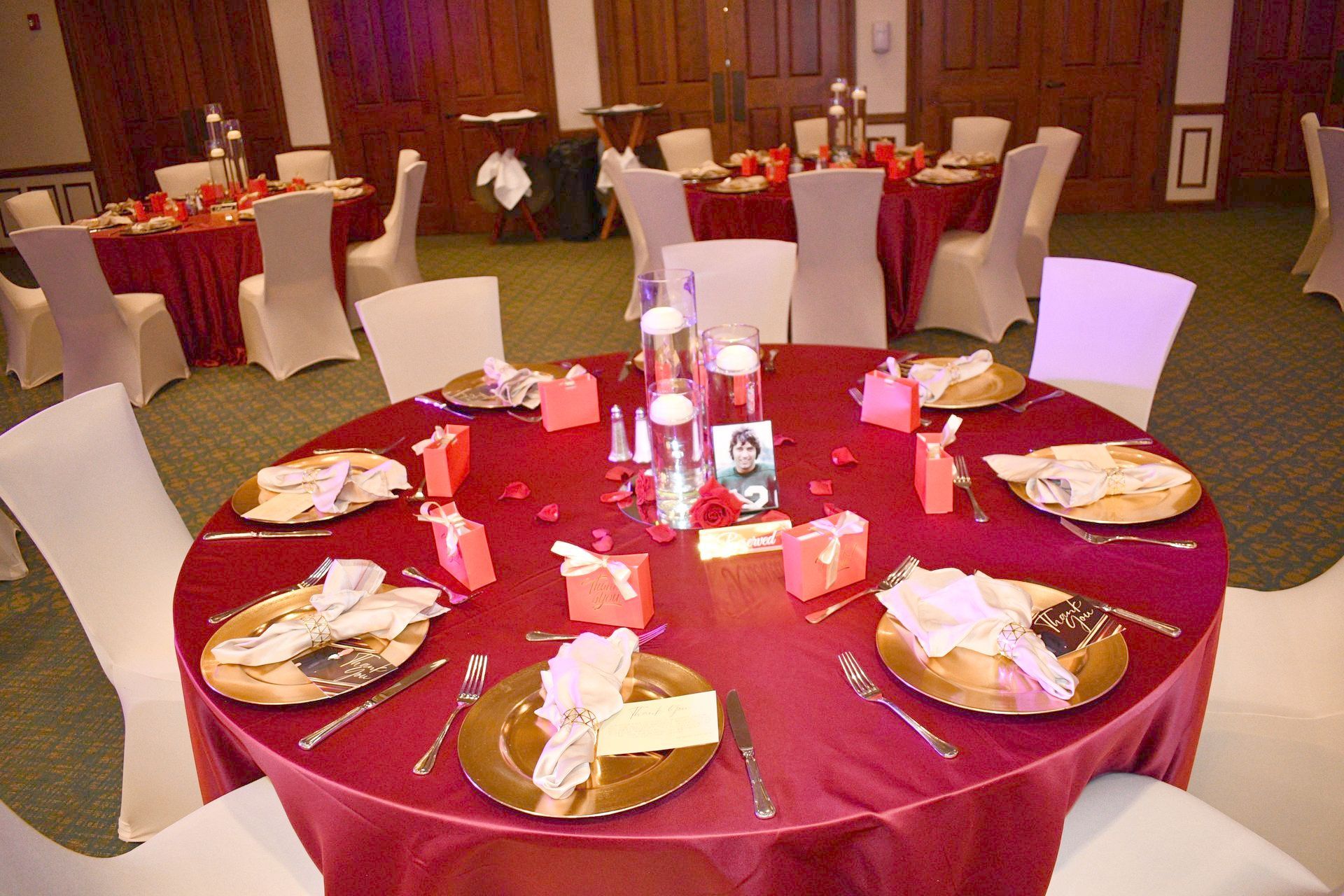 Round tables with red tablecloths, set for a formal event. White chairs, gold chargers, and centerpieces are visible.