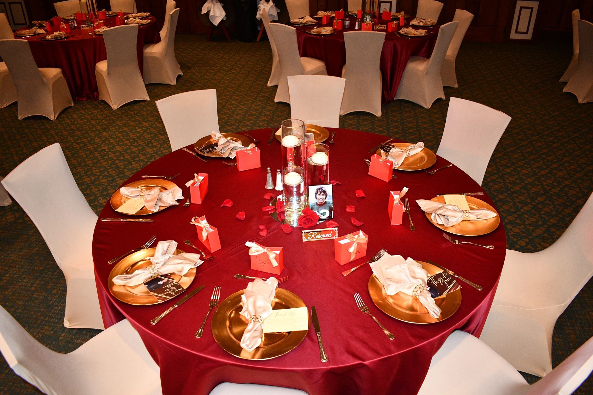 Round table set for a formal event with red tablecloth, gold plates, white chairs.