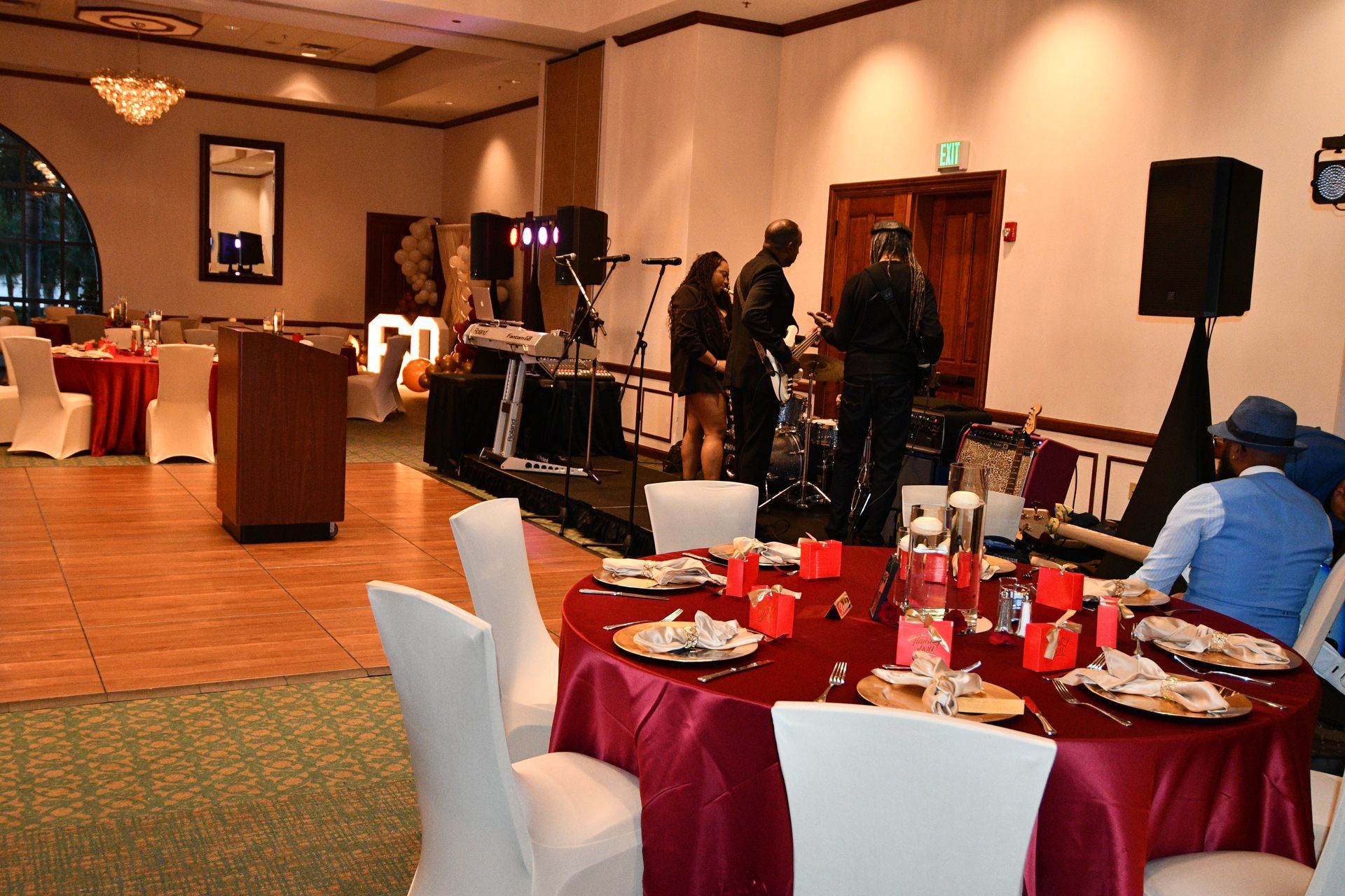 Band playing on stage in banquet hall. Round tables set with red cloths, guests seated.