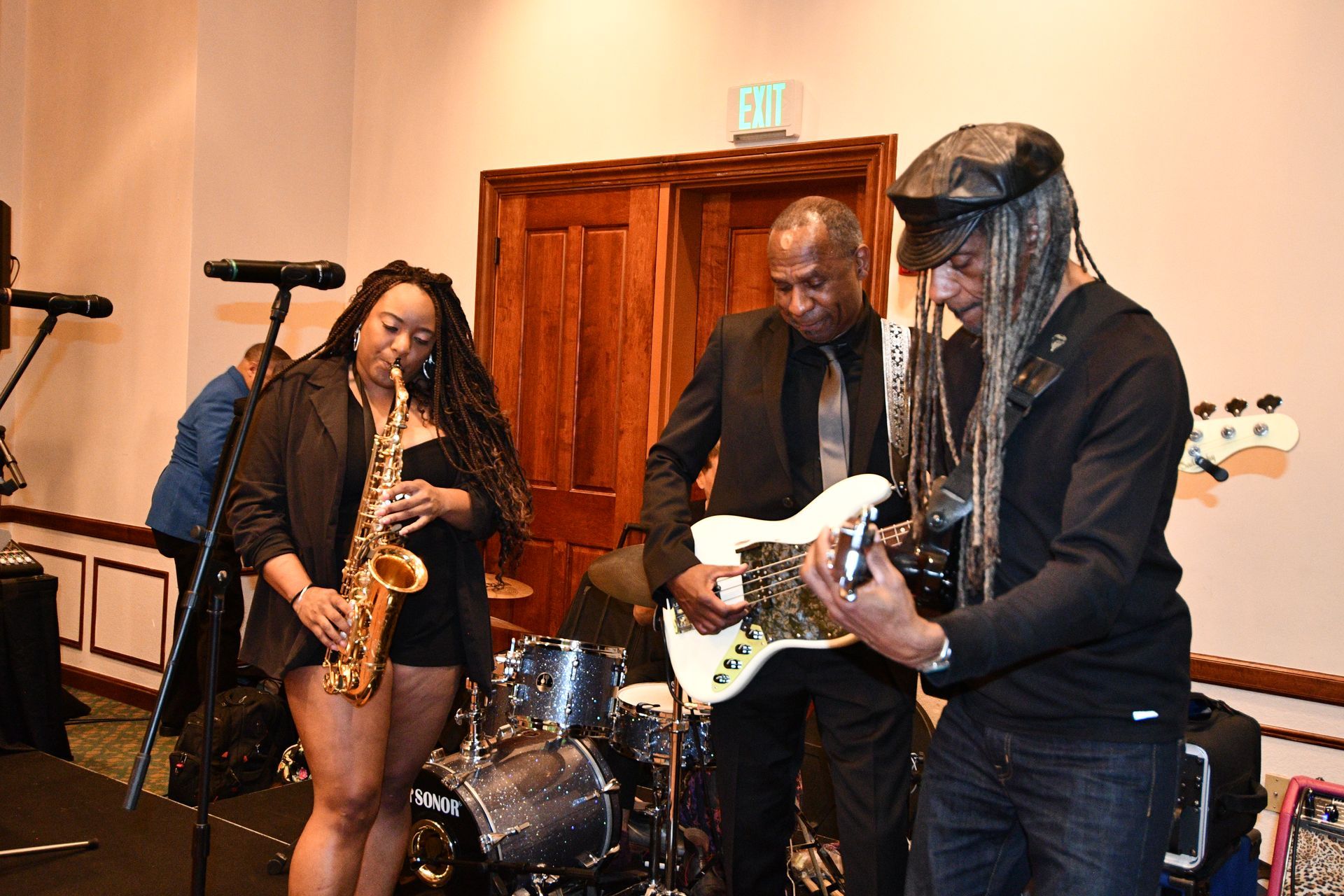 Band performing on stage: Saxophonist, guitarist, bassist, and drummer. All are African American, performing live.