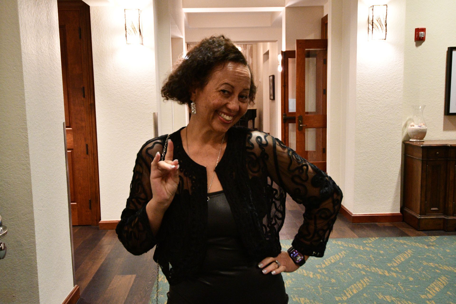 Woman in black outfit, smiling, making peace sign in hallway.