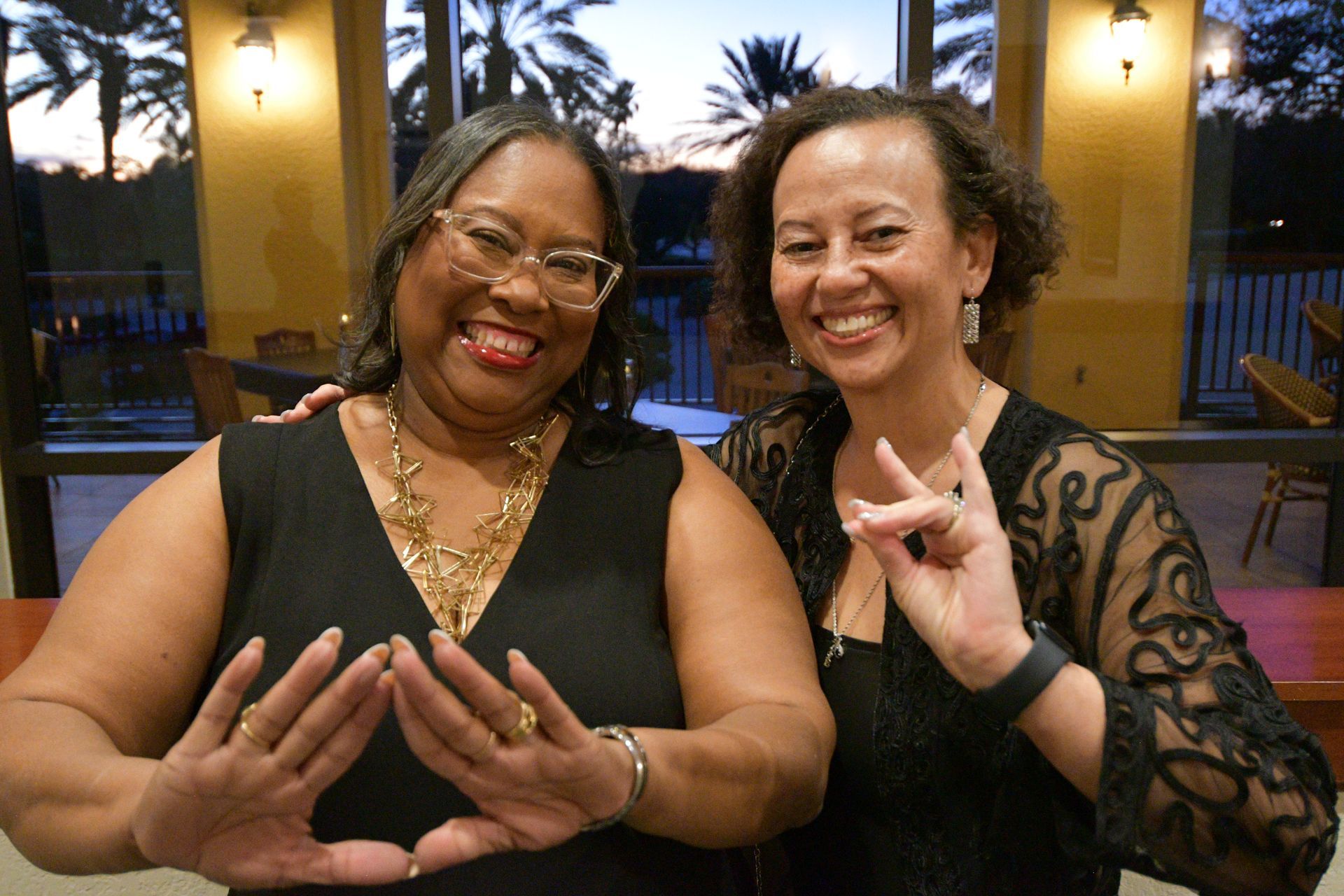 Two women smiling, posing, hands forming a shape, in a brightly lit room with a view of an outside area.