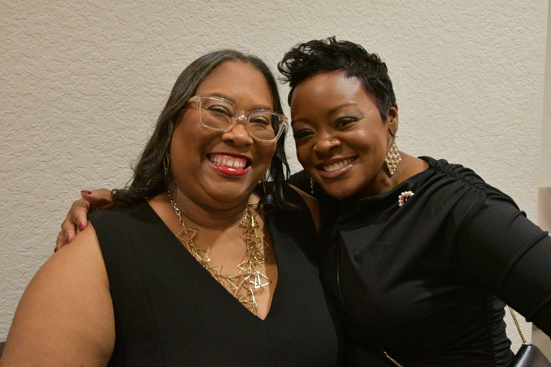 Two smiling people in black outfits, indoors. One has long hair, glasses, and a necklace. The other has short hair.