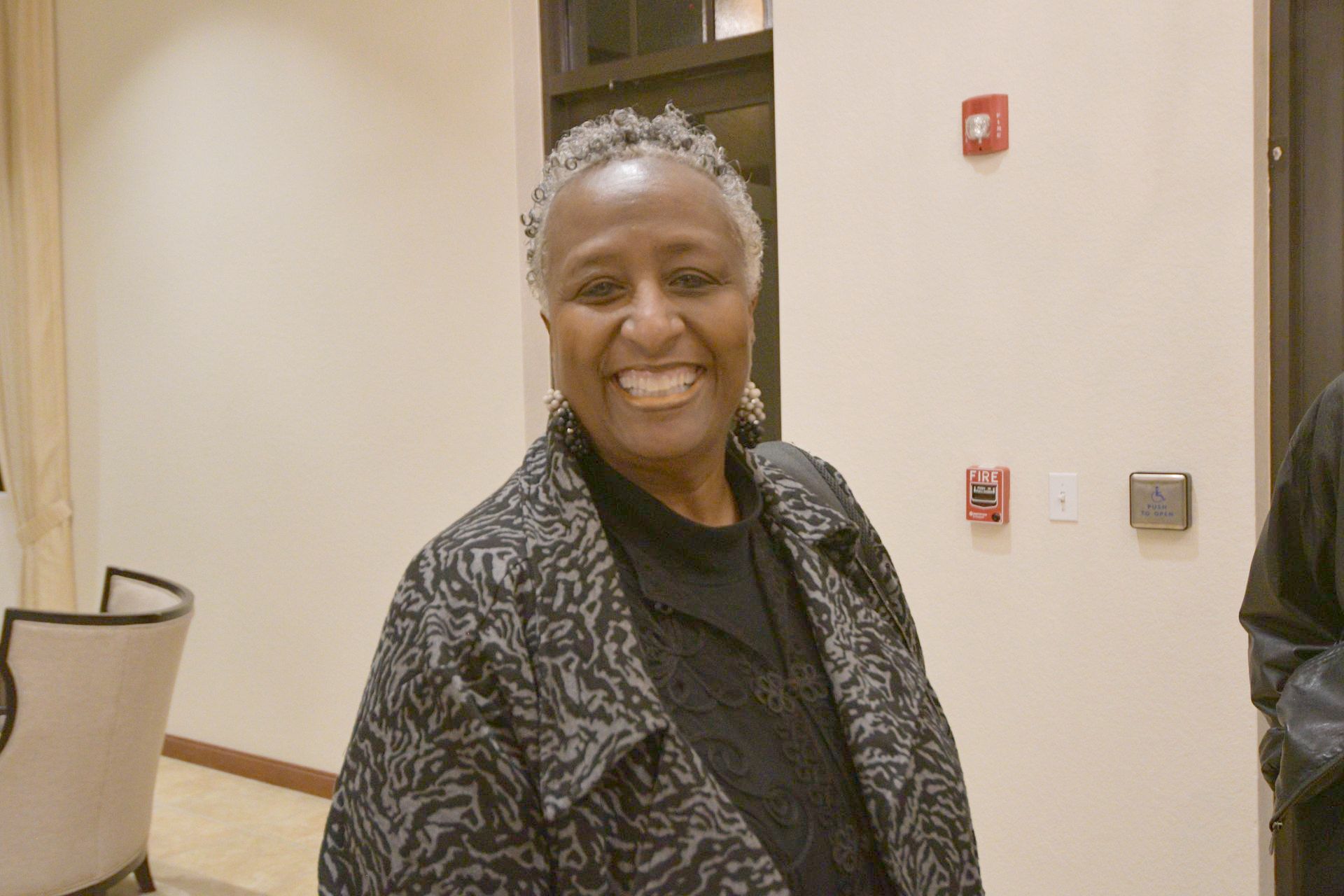 Woman with gray hair and patterned jacket smiles indoors near a wall with a fire alarm.