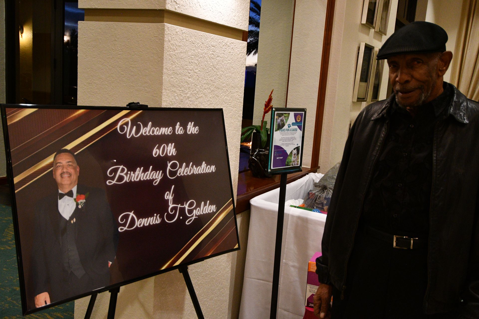 Man standing near a sign for a 60th birthday celebration. Sign features a photo of a man in a tuxedo.