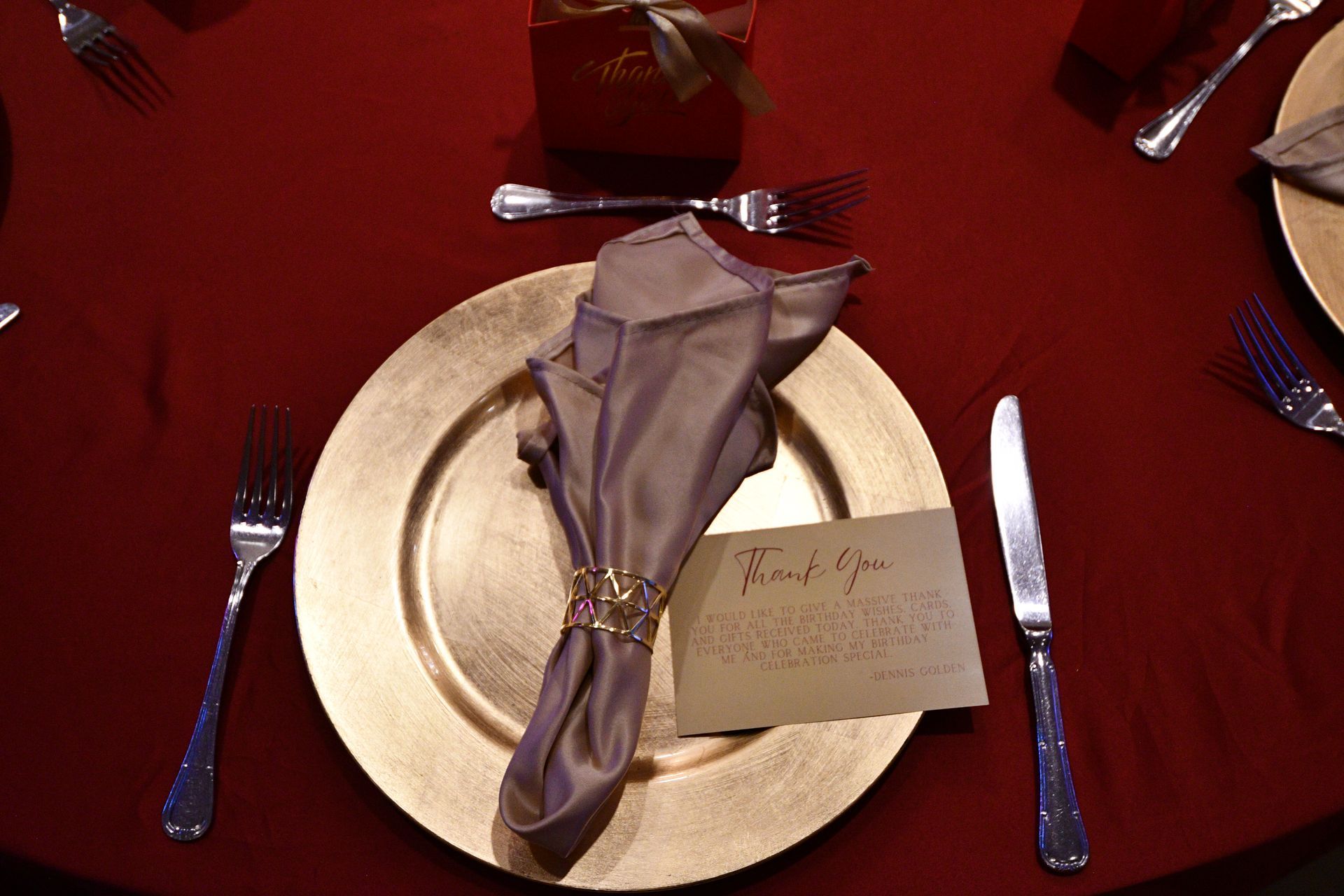 Place setting with gold charger, folded napkin, silverware, and thank you card on red tablecloth.