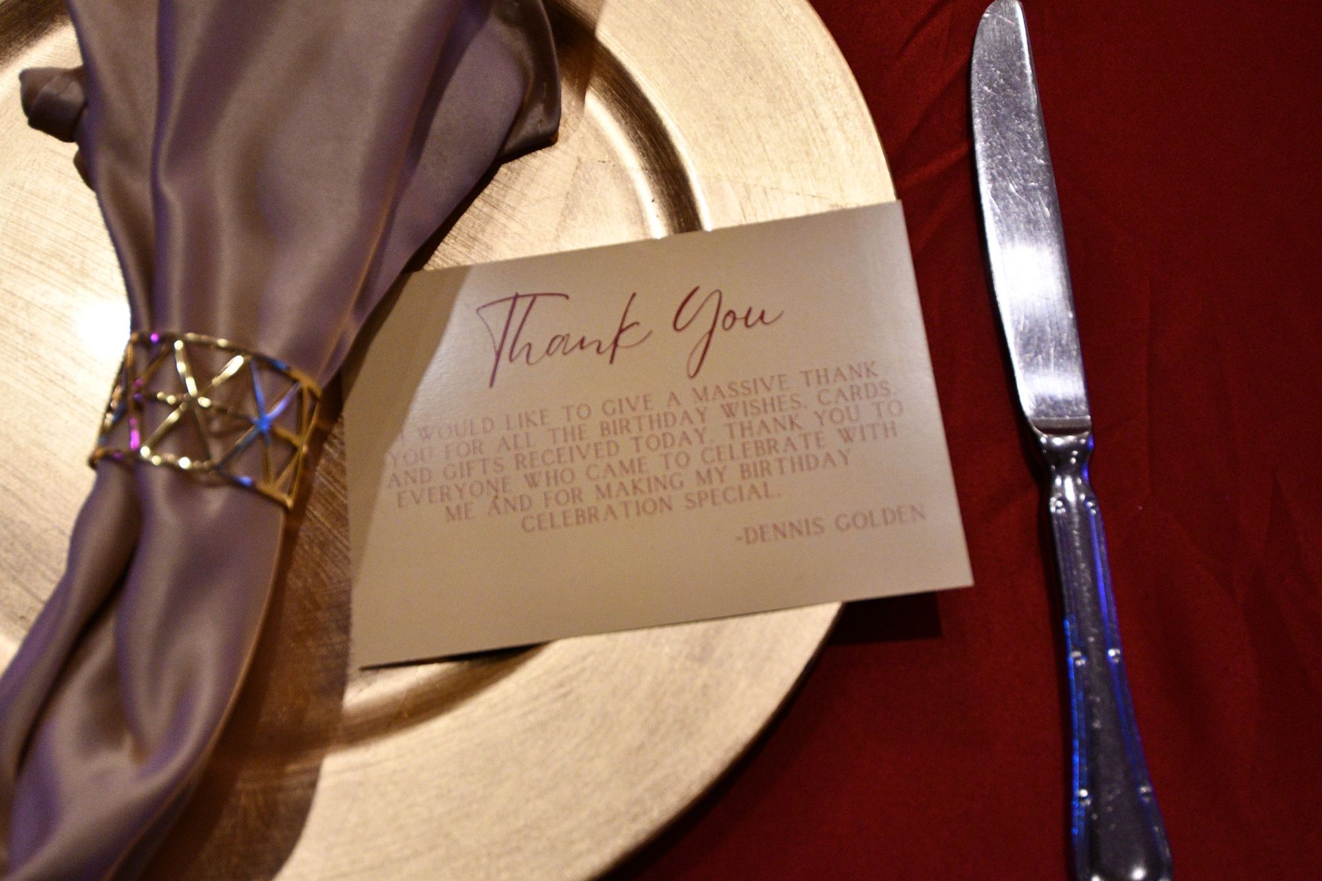Place setting with gold charger, napkin, thank you card, and knife. Napkin held by a gold ring.