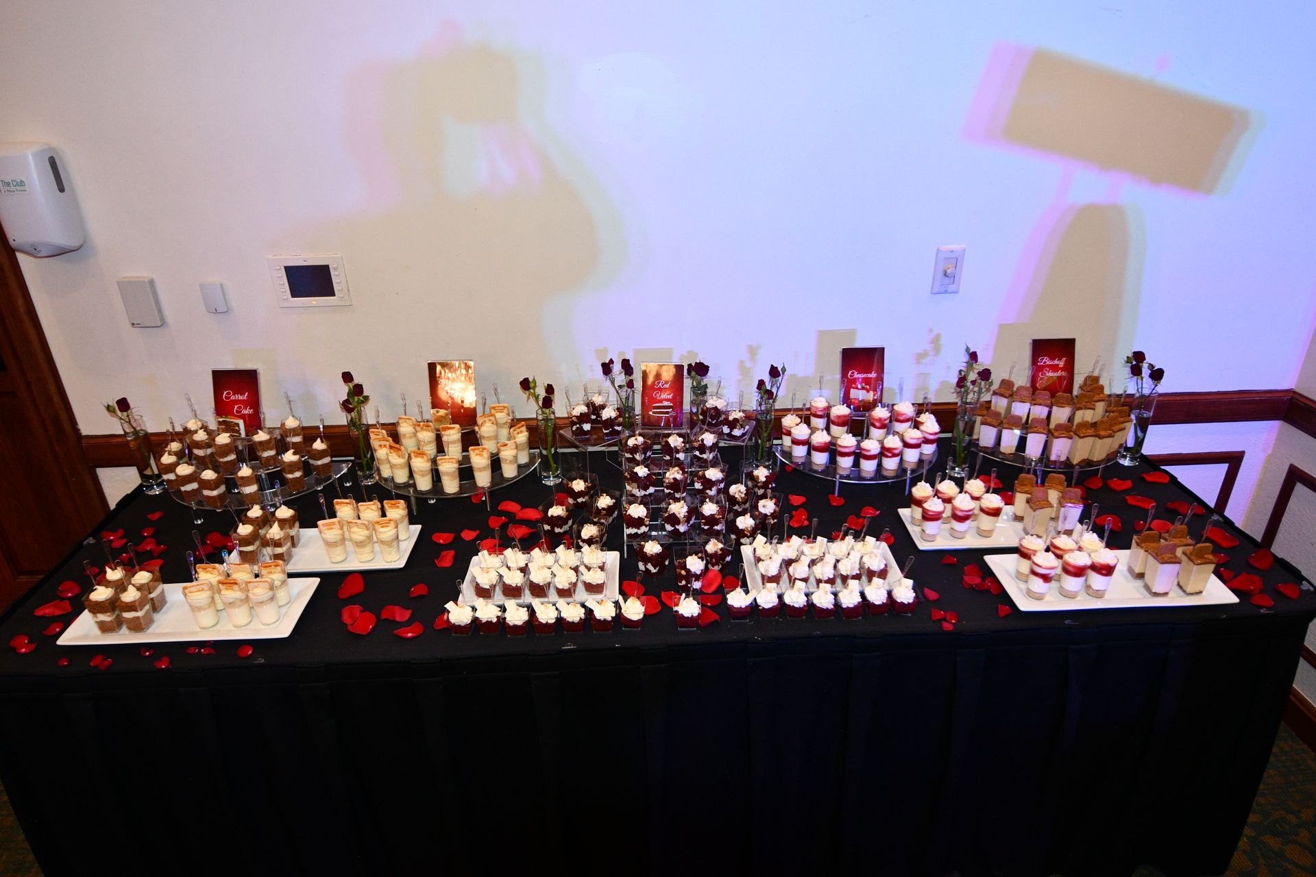 Dessert buffet with various small treats, arranged on a black tablecloth. Red rose petals are scattered on the table.