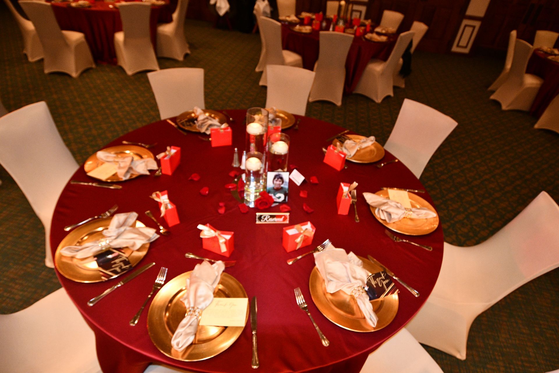 Round table set for a formal event with red tablecloth, gold chargers, white chairs, and centerpieces.
