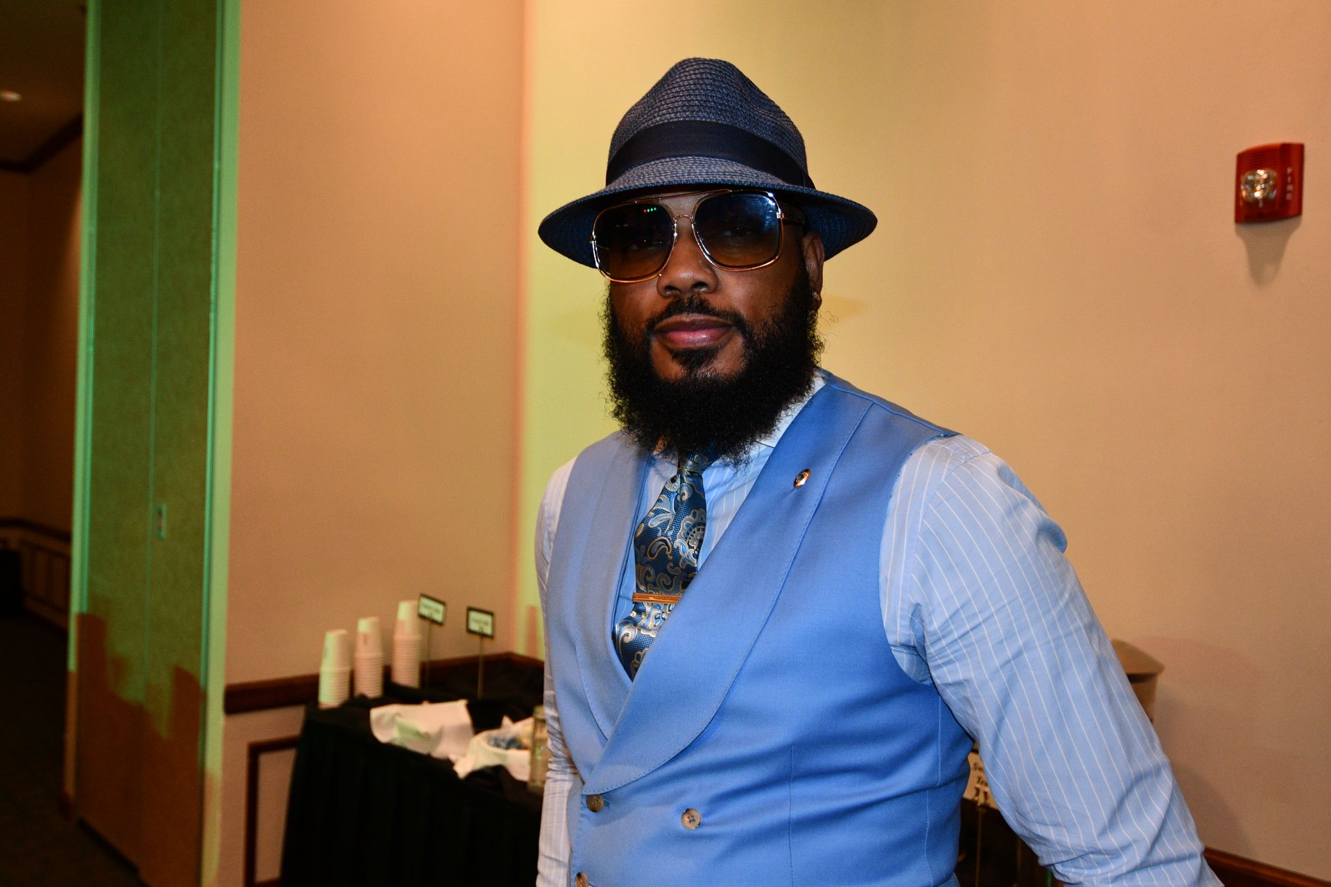 Man in blue suit, hat, and sunglasses, standing near a wall in a room.