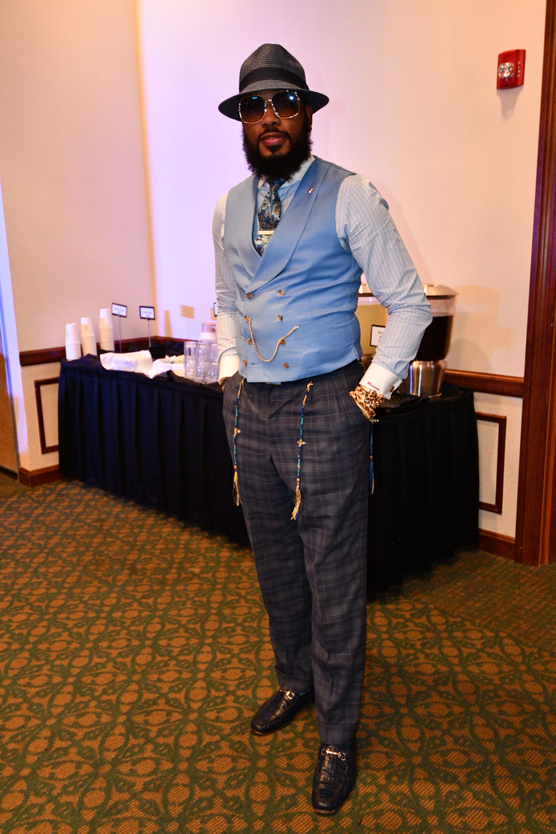 Man in stylish attire, hat, vest, and patterned pants, posing indoors with a food service table in the background.