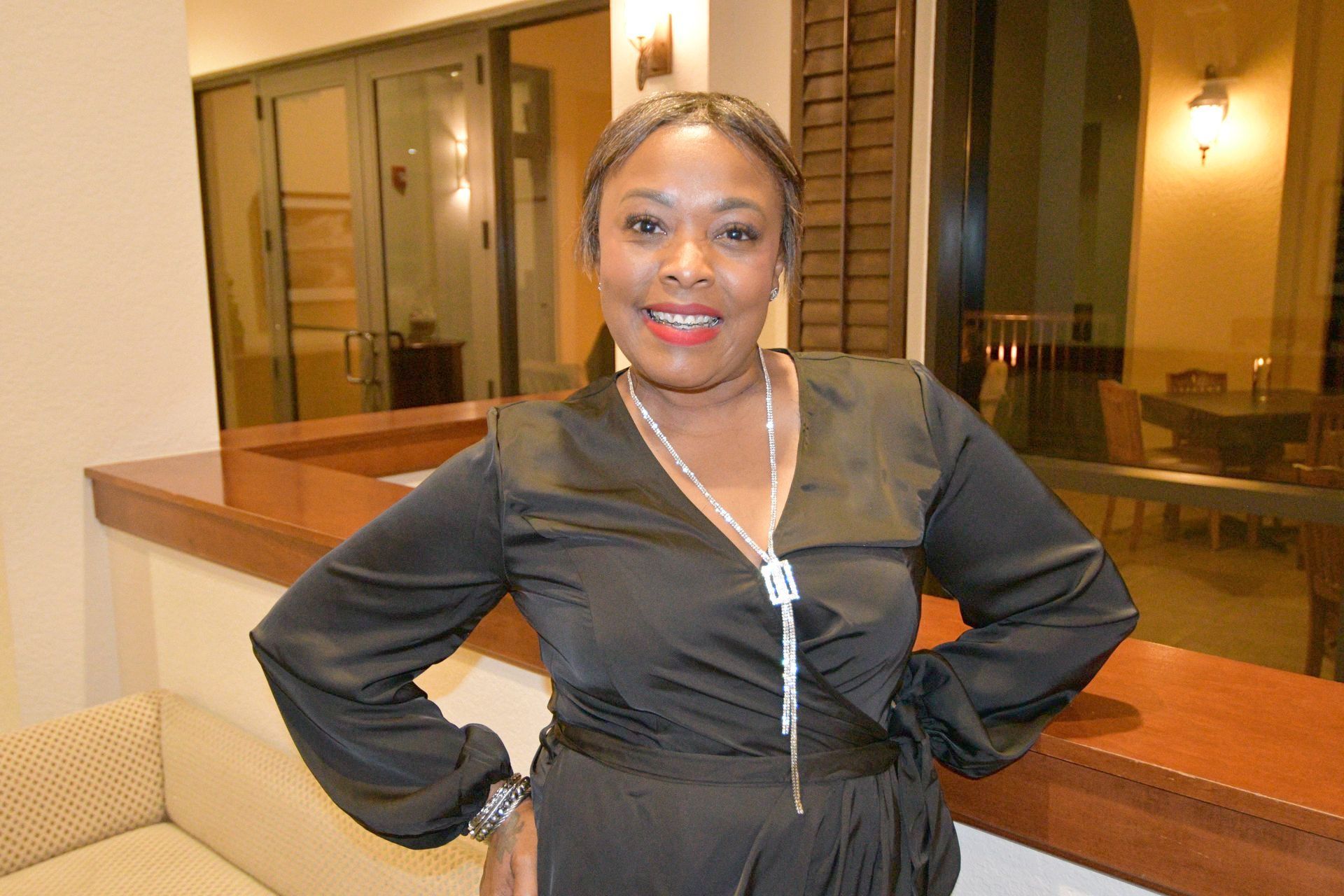 Woman in a black dress with silver necklace, smiling. Standing by a window, arms on hips.