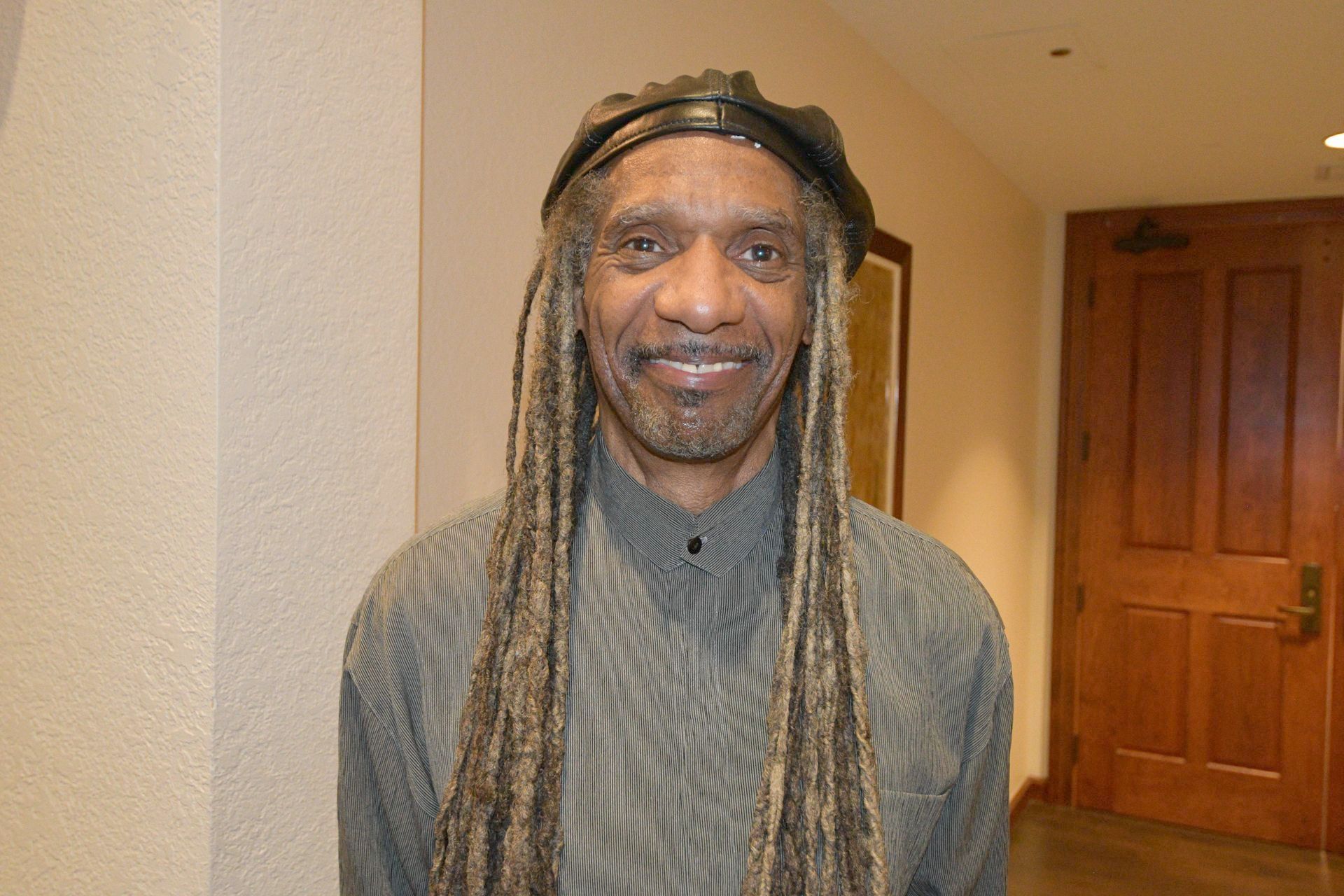 Man with long dreadlocks and a hat smiles, standing near a wall and a wooden door.