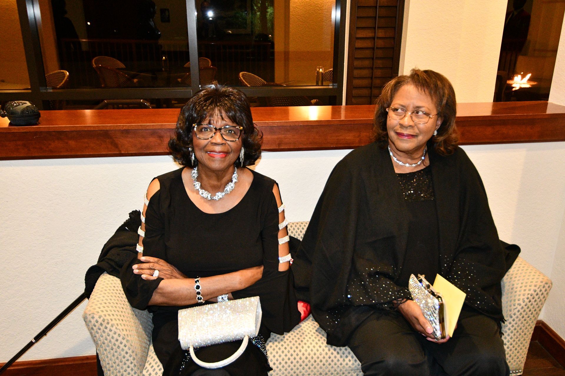 Two women sit on a beige sofa. One smiles with arms crossed, holding a purse. The other holds a notebook.