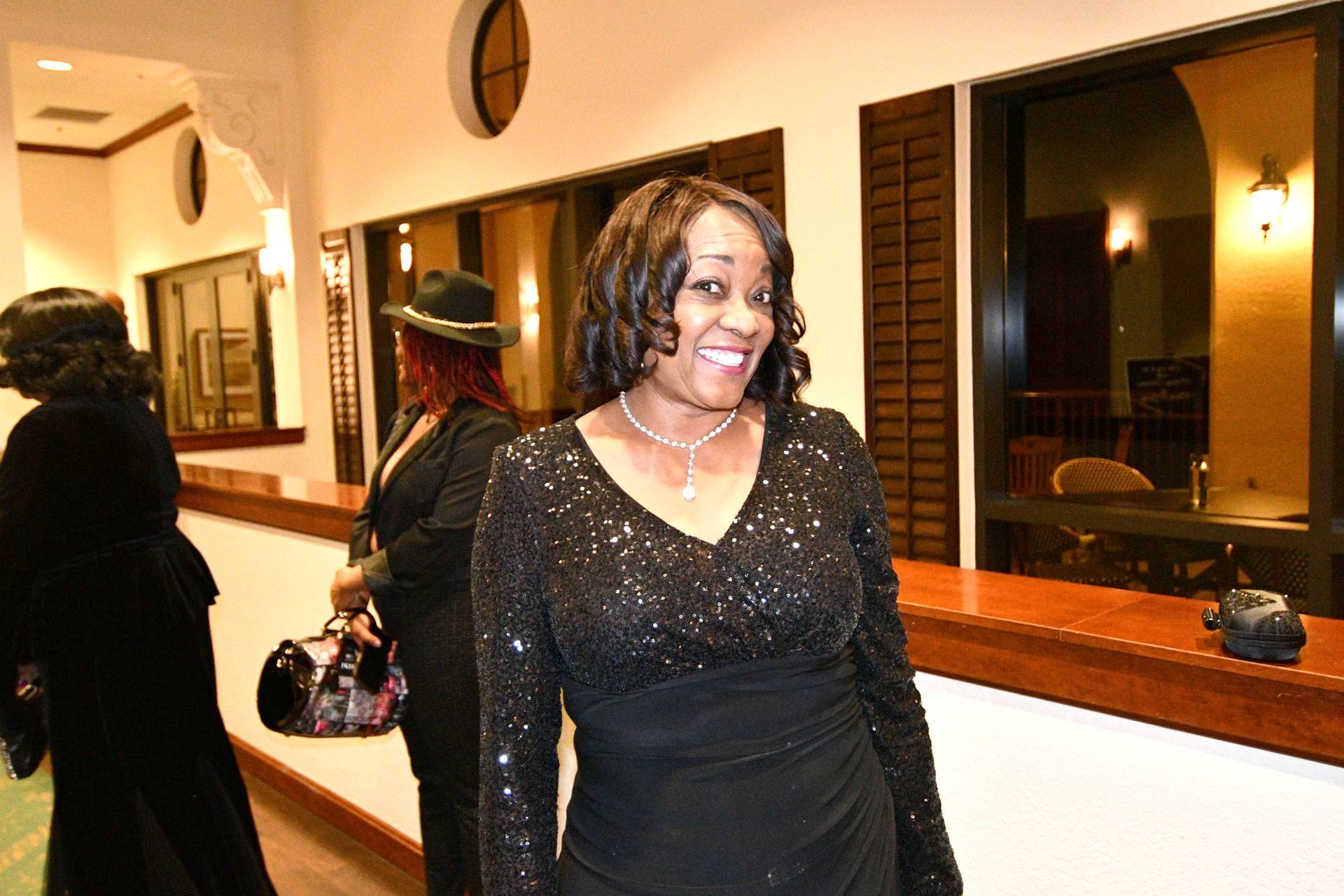 Woman in sparkly black dress smiles, standing near a wooden counter, with two other people in the background.