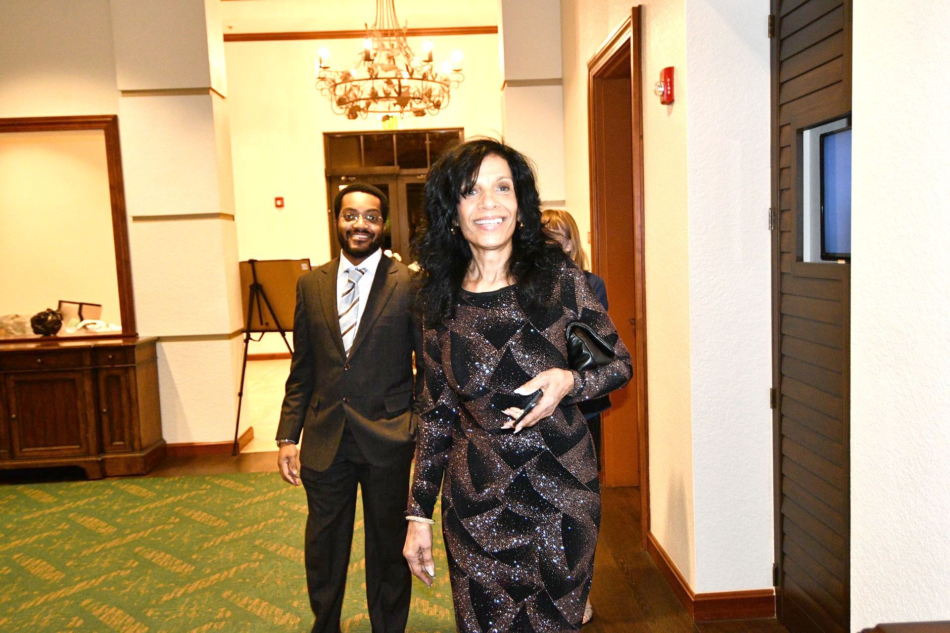 Woman in sequined dress and man in suit walking in hall; chandelier in background.