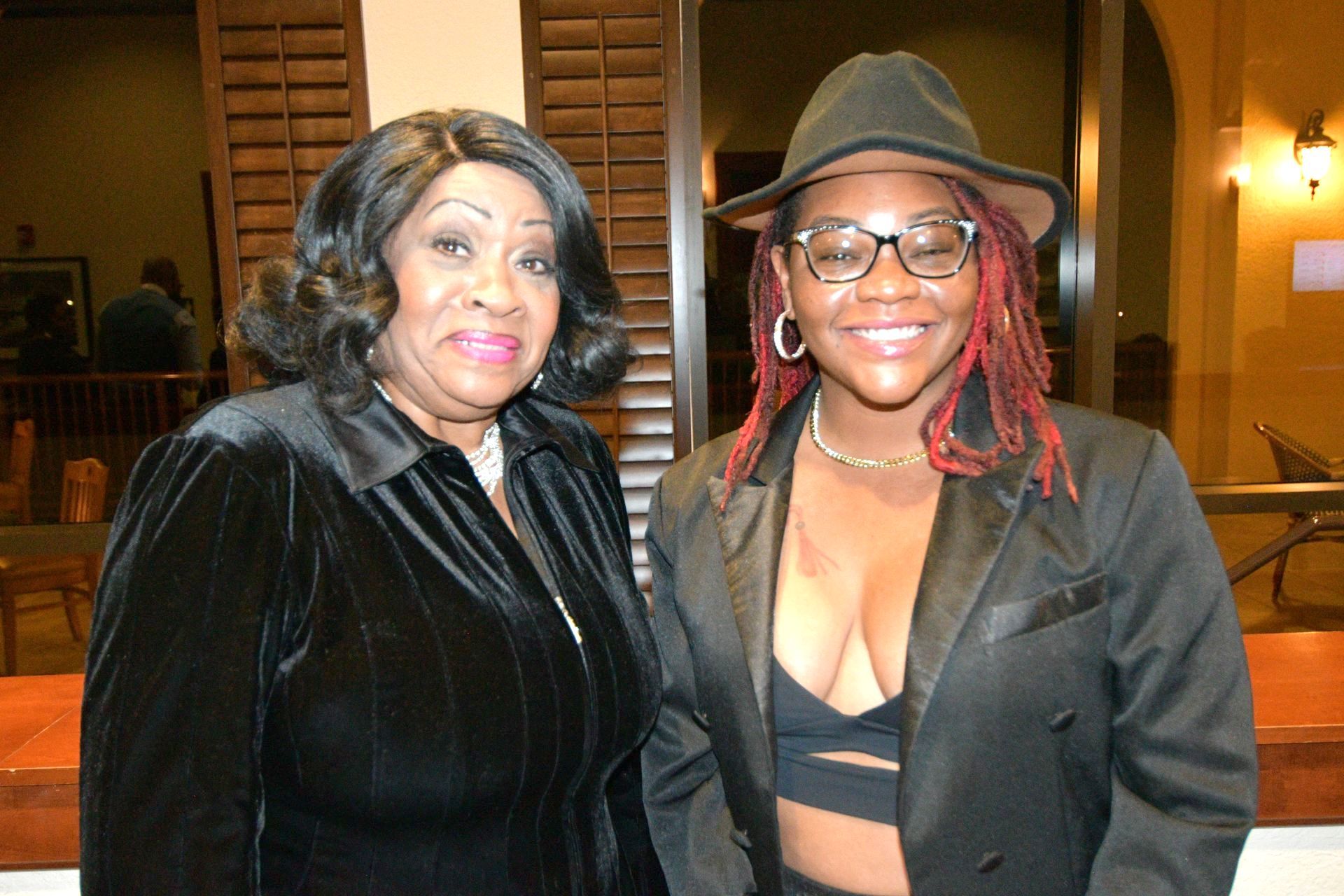 Two women smiling, posing indoors. One wears a black jacket, hat, and glasses; the other a black suit and has wavy hair.