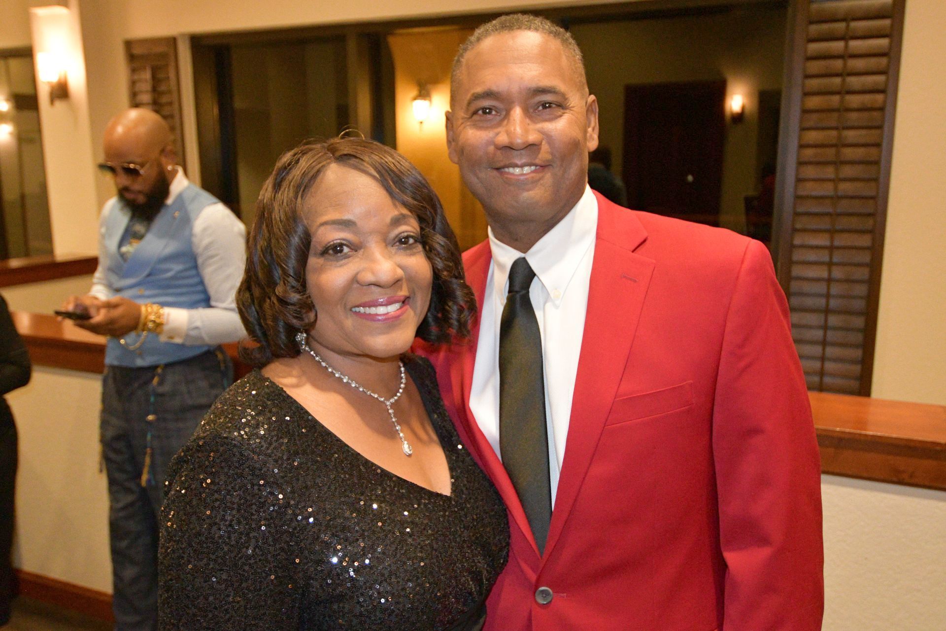 Couple smiling at the camera, man in red suit, woman in black sequin dress, blurred figure in background.