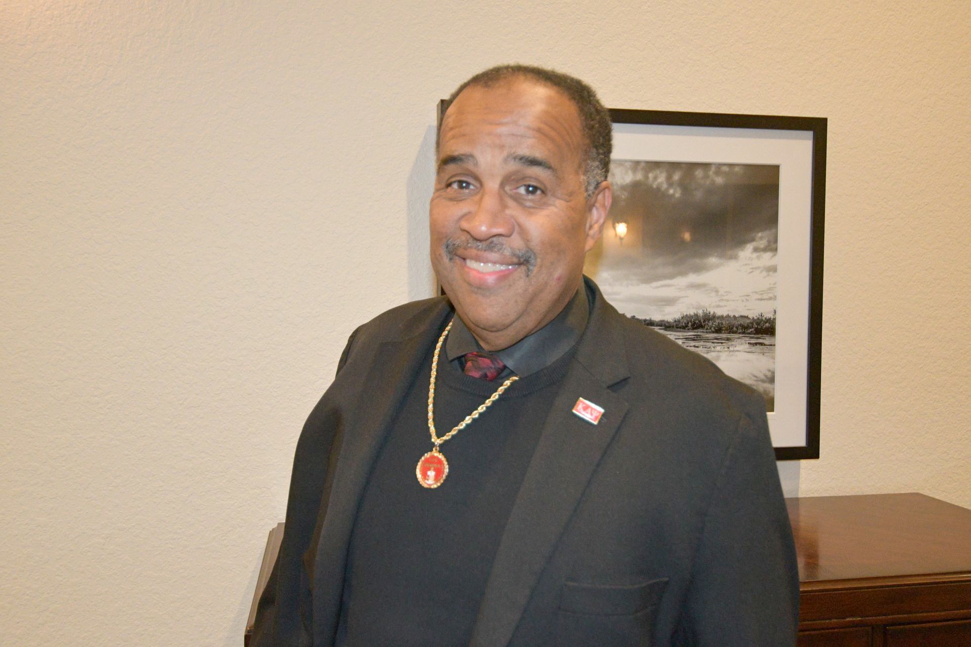 Man in a suit smiles, wearing a necklace. He stands indoors near a framed photo.
