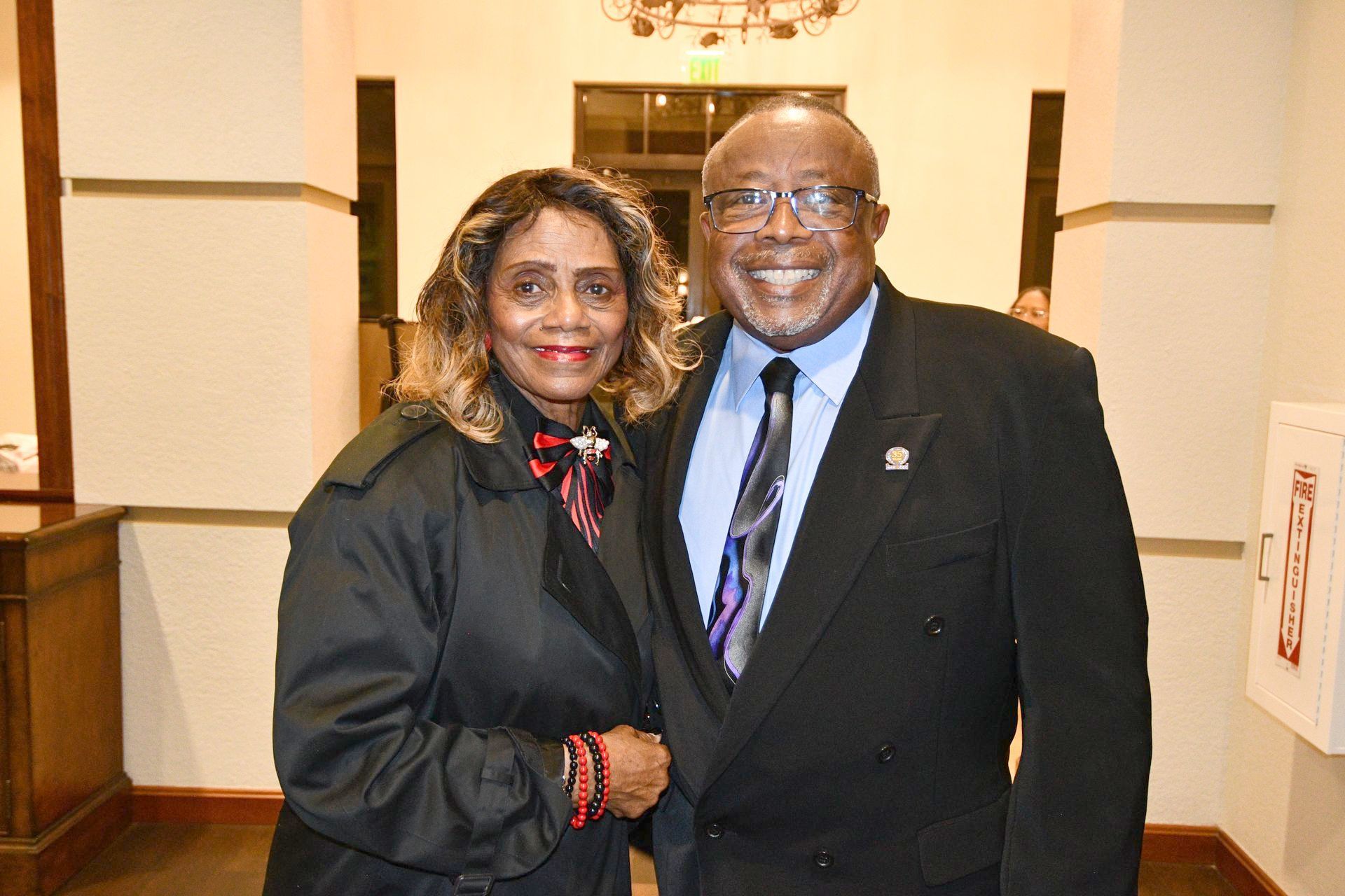 A smiling man and woman pose together indoors. The man wears a suit and tie, and the woman wears a coat and scarf.