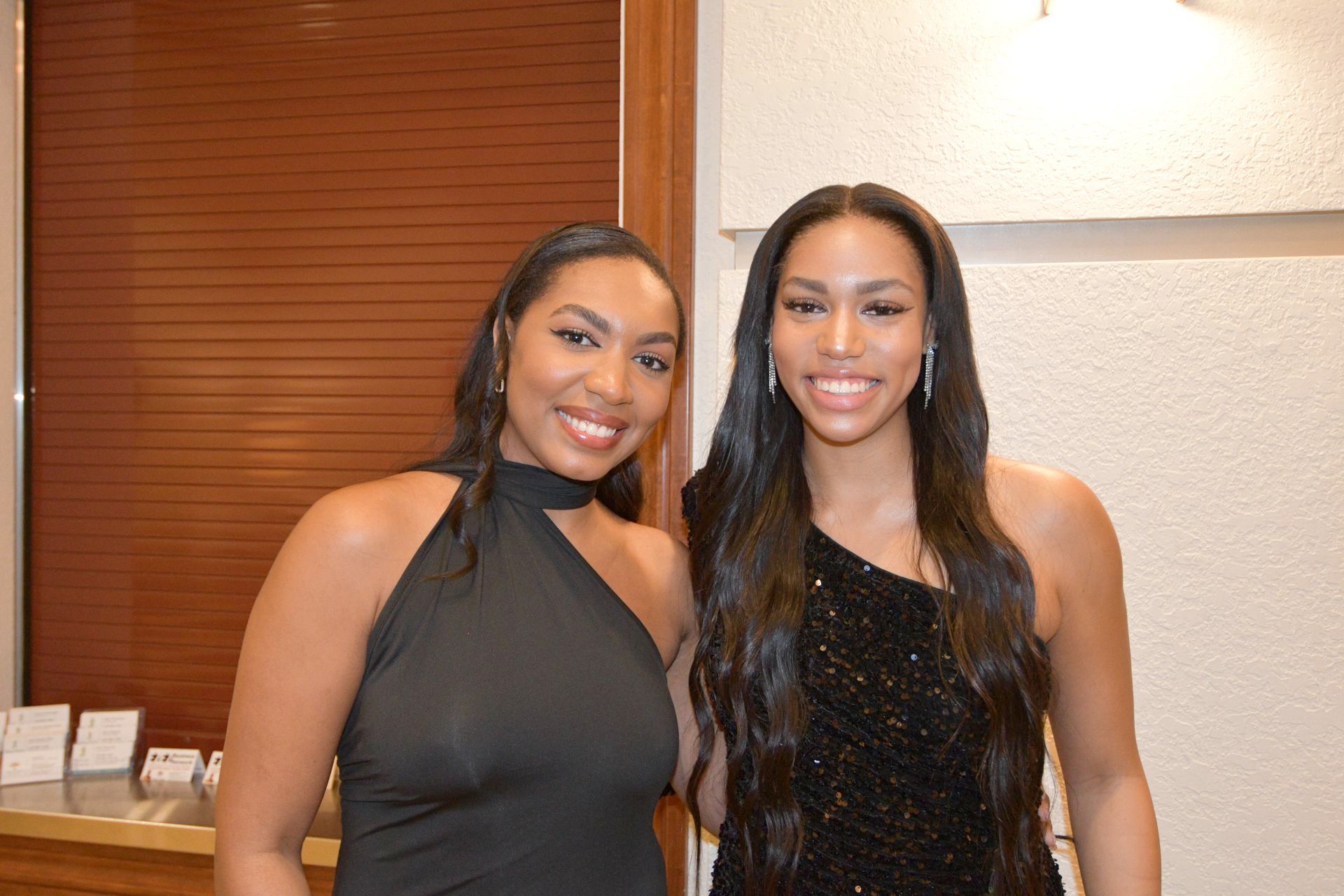 Two women smiling, posing for a photo. One wears a black halter dress, the other a sequined one-shoulder dress. Indoors.