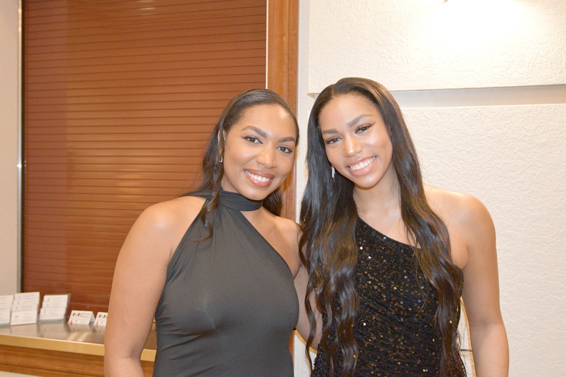 Two women smiling, posing for a photo. One in a black halter dress, other in a one-shoulder sequin gown. Indoors.