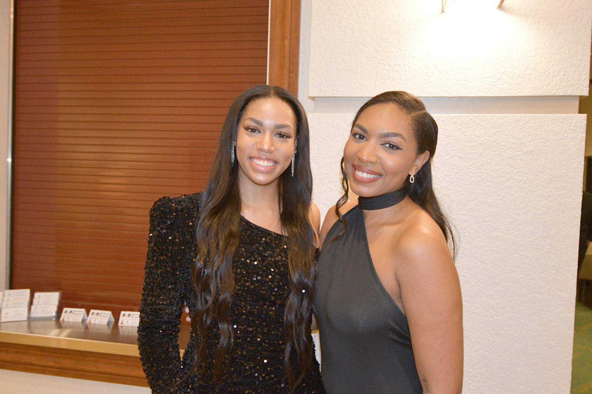 Two women smiling, posing for a photo. One in a black, sequined dress; the other in a black halter dress.